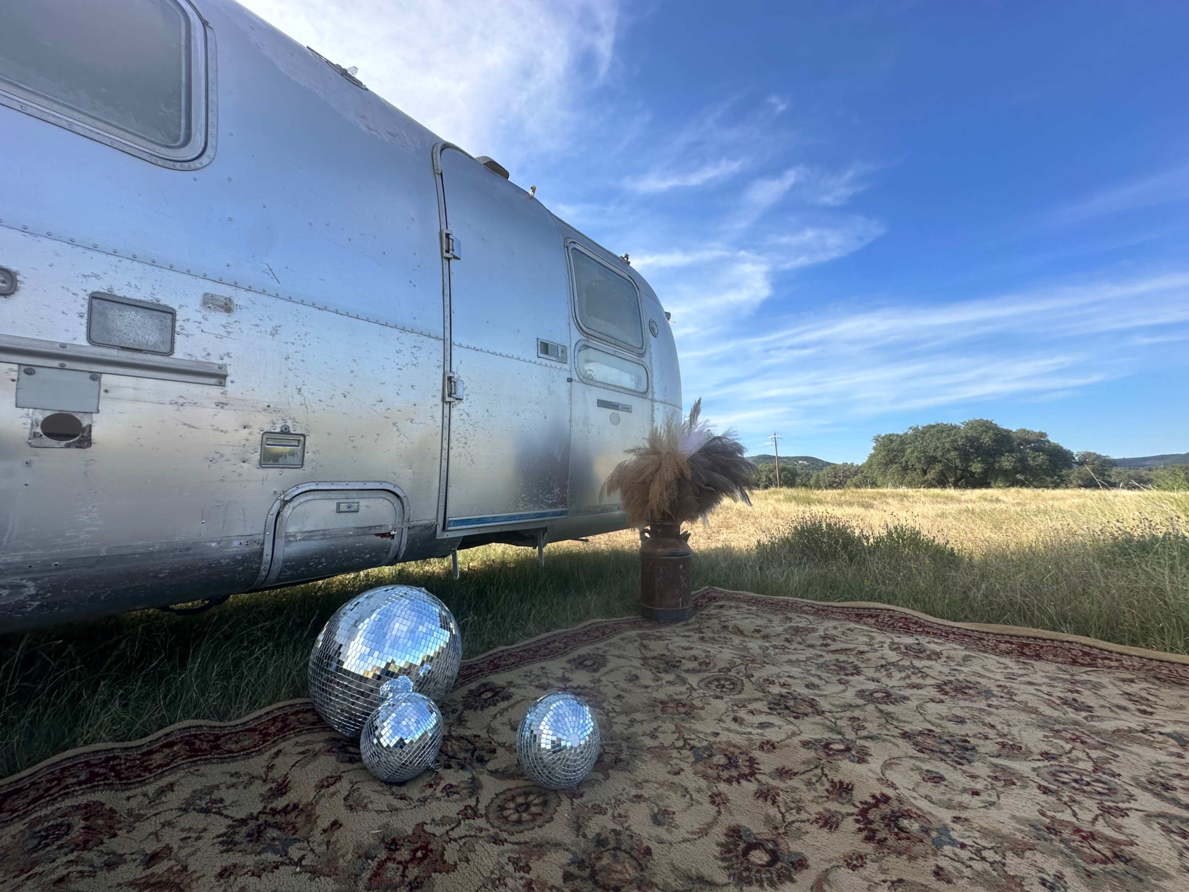 An Airstream trailer is parked beside a rug, with three disco balls and a decorative plant nearby, set against a backdrop of a grassy field and blue sky.