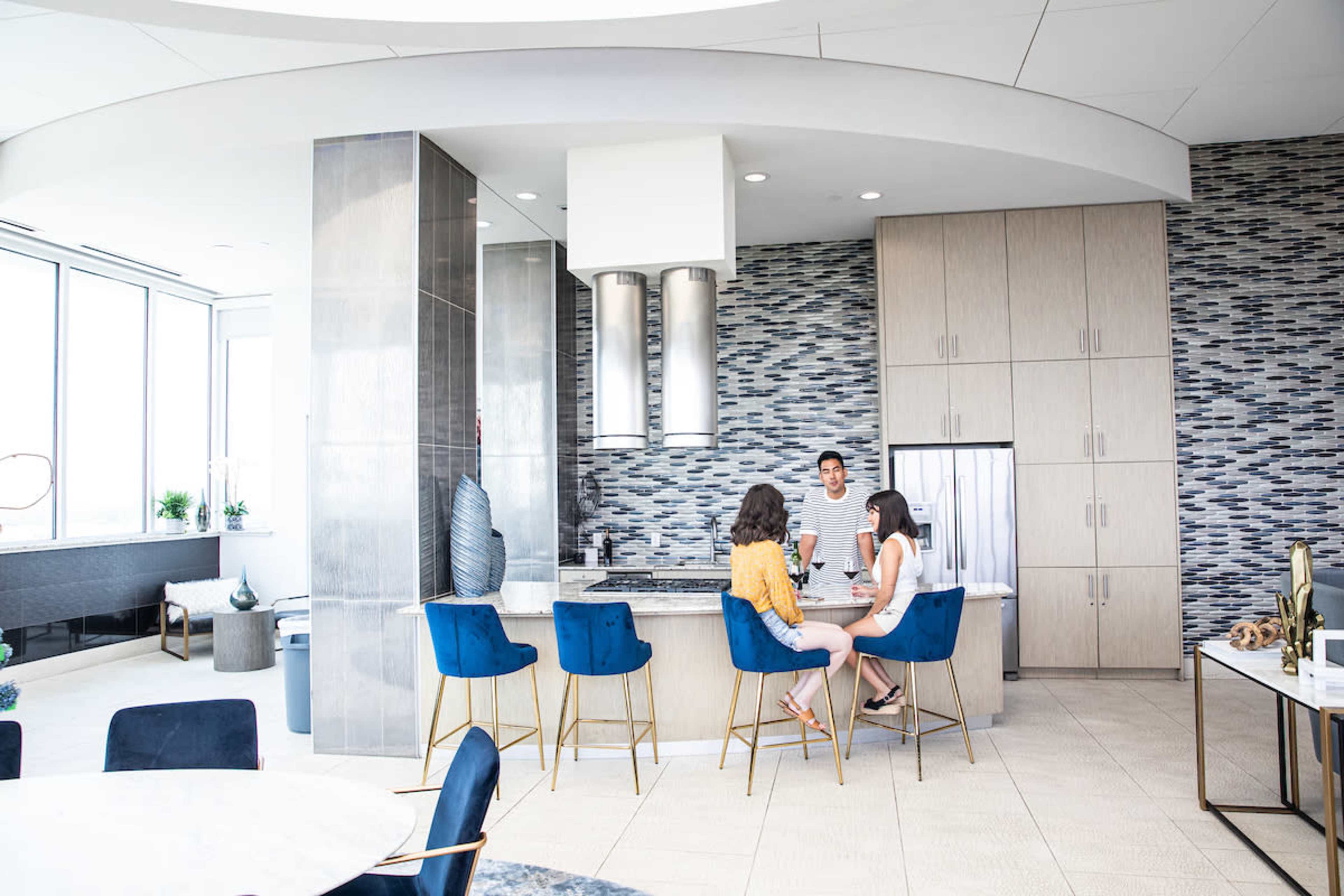 A modern kitchen features a central island with three stools as two women engage in conversation, while a man stands nearby by the cooking area.