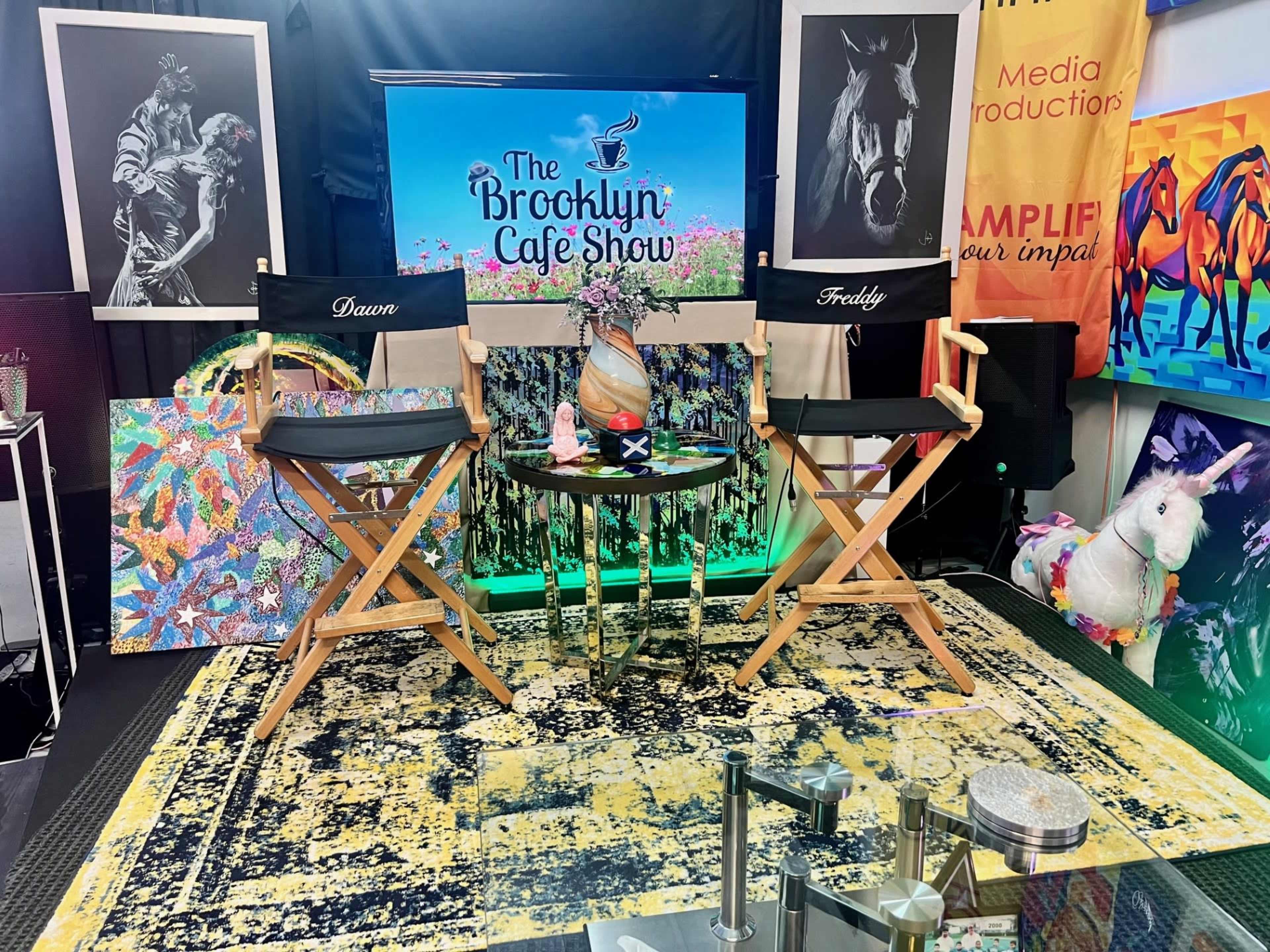 A studio setup for "The Brooklyn Cafe Show," featuring two director's chairs labeled "Dawn" and "Freddy," surrounded by colorful artwork and a backdrop with the show's logo.