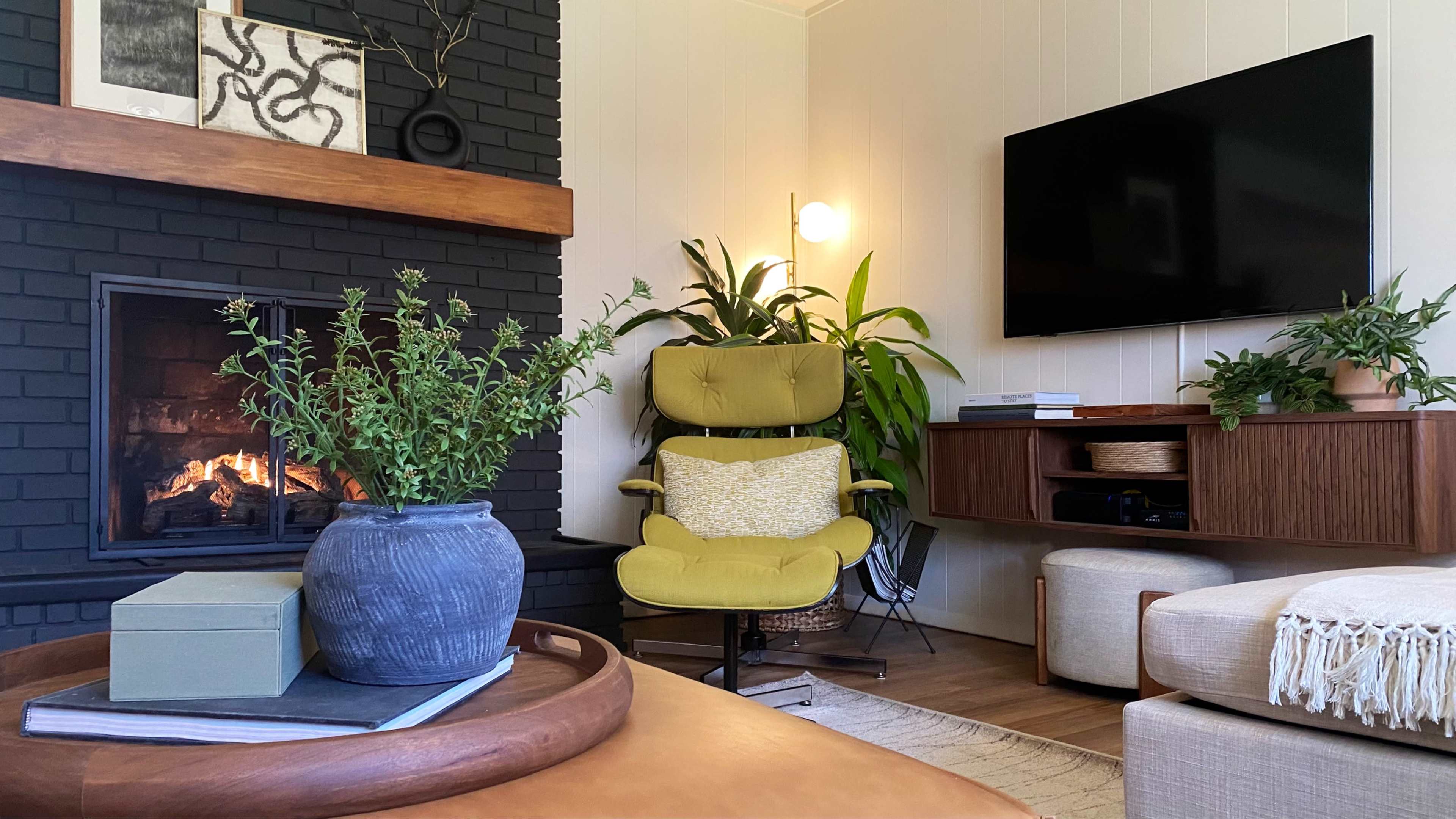 A cozy living room features a green armchair, a modern TV mounted on the wall, and a fireplace, surrounded by various plants and decorative elements.