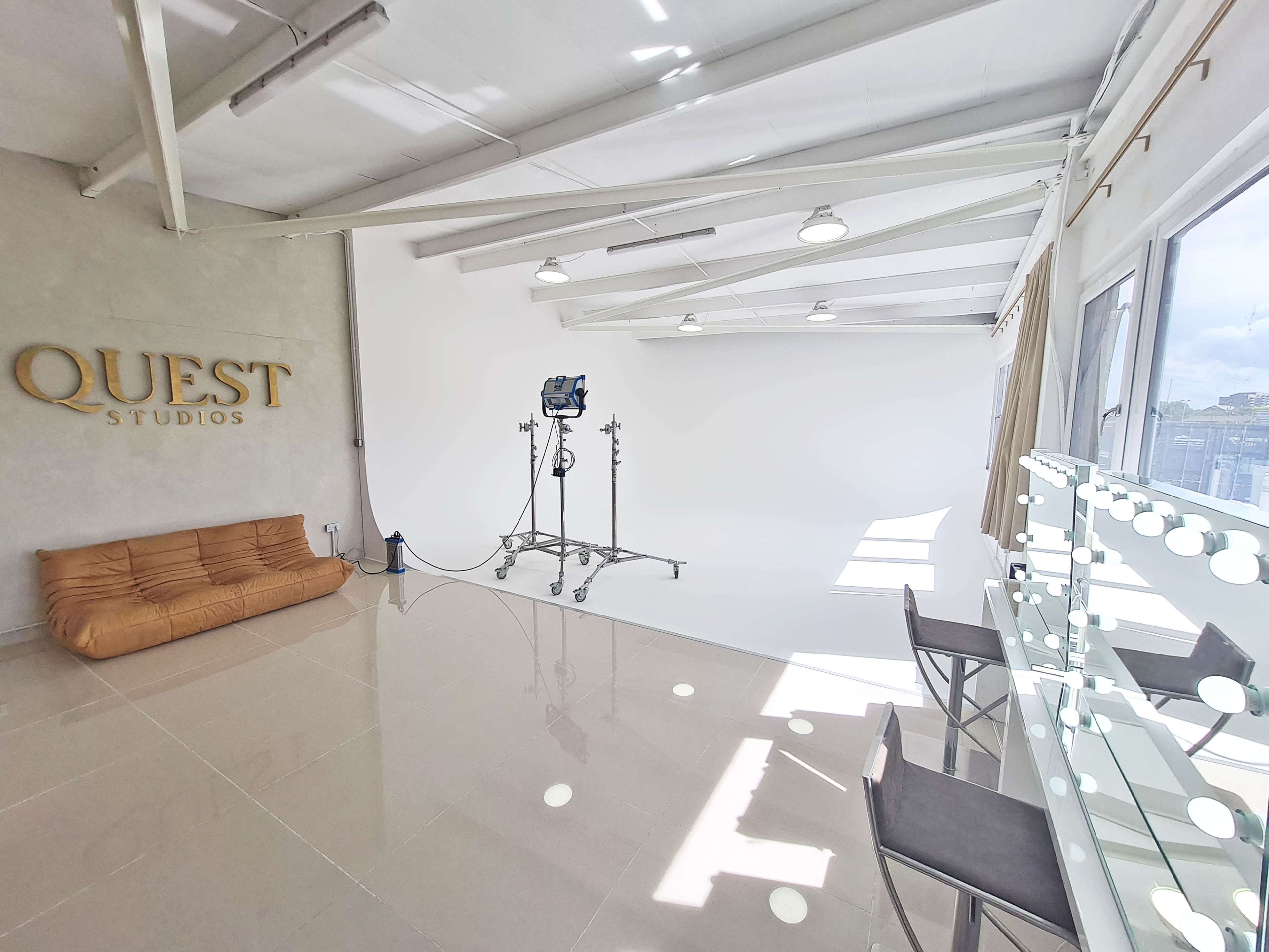 A spacious studio features a beige couch, a lighting setup on a stand, and a vanity with illuminated mirrors, all against a white backdrop.