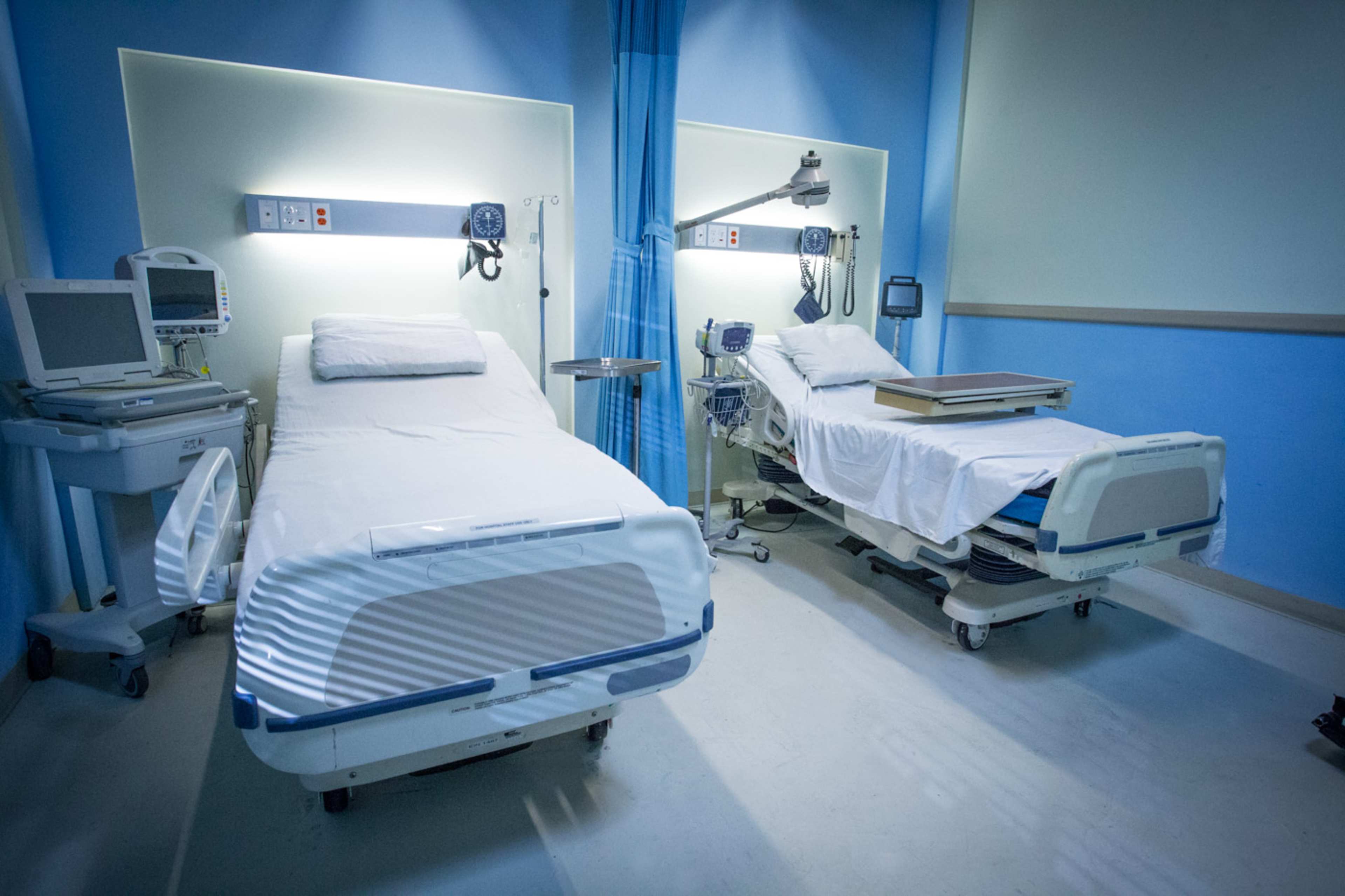 A hospital room with two empty beds positioned alongside medical equipment and monitors against light blue walls.