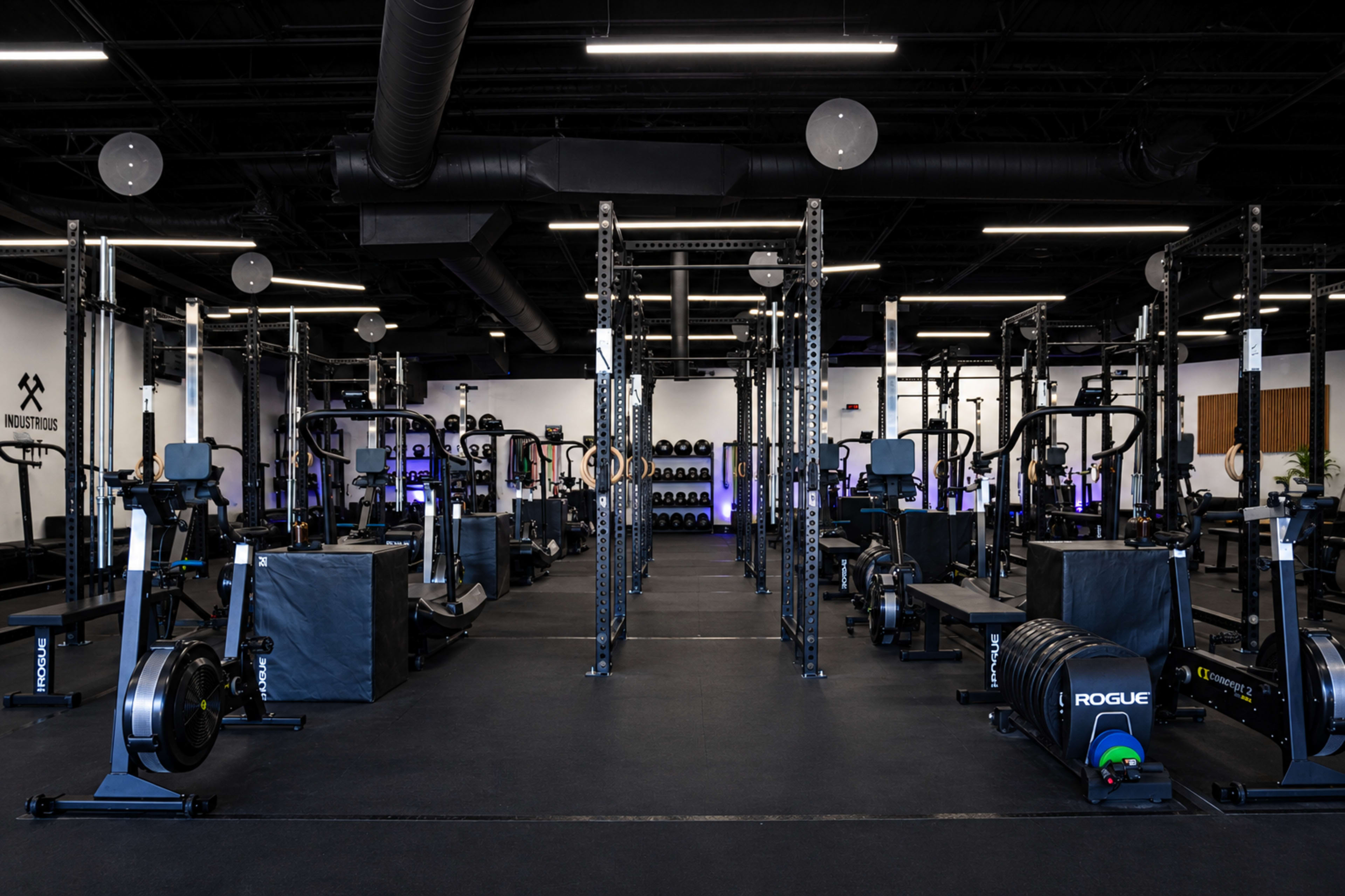 The image shows a spacious gym filled with various workout equipment, including rowing machines, weightlifting racks, and free weights, arranged in an organized layout.