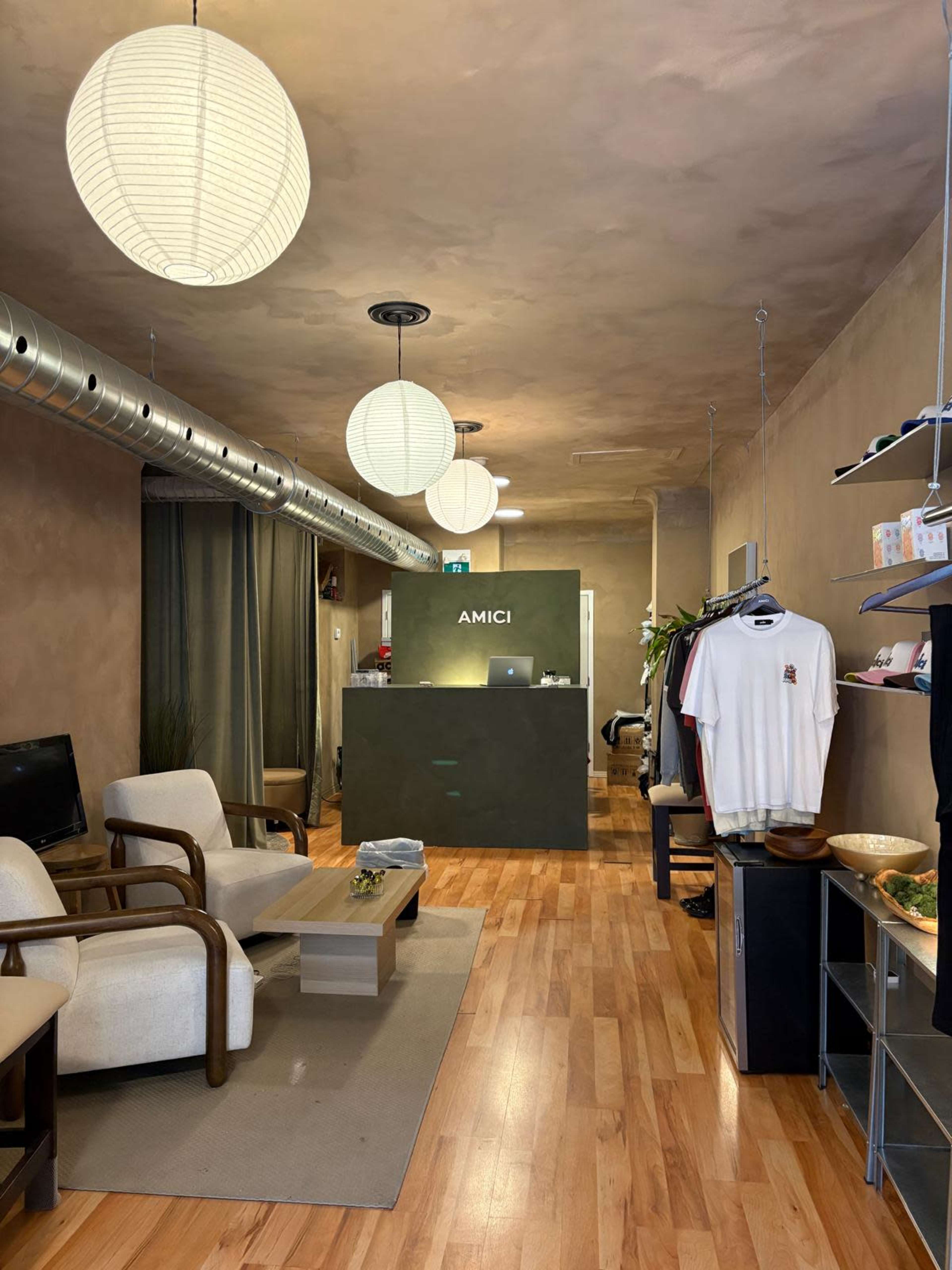 The image shows a modern retail space with wooden flooring, a reception area marked "AMICI," and clothing displayed on racks along the walls.