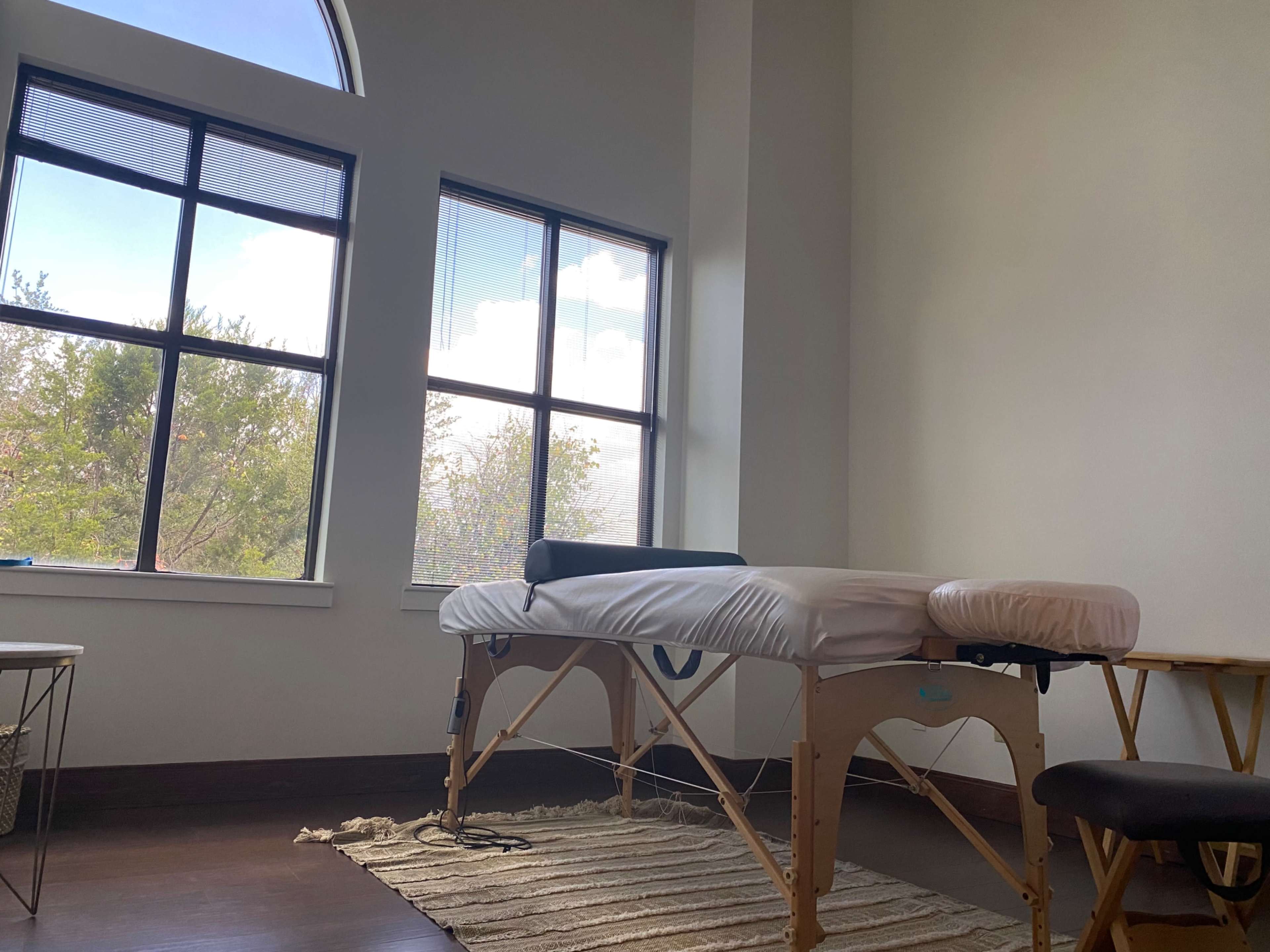 The image shows a massage table positioned in a room with large windows and natural light filtering through.