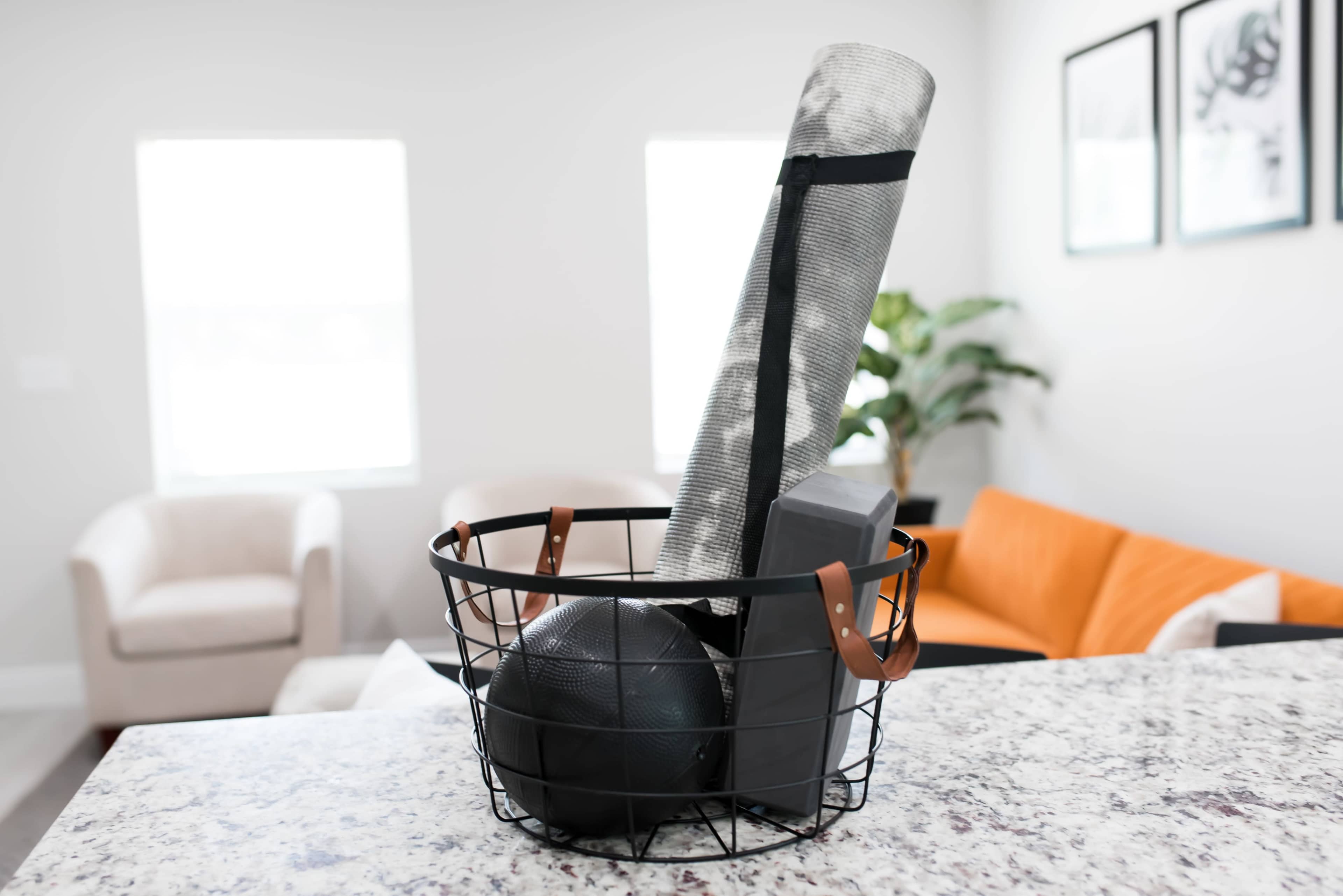 A wire basket on a granite countertop holds a yoga mat, a dumbbell, and a basketball, set against a brightly lit living area with modern furniture.