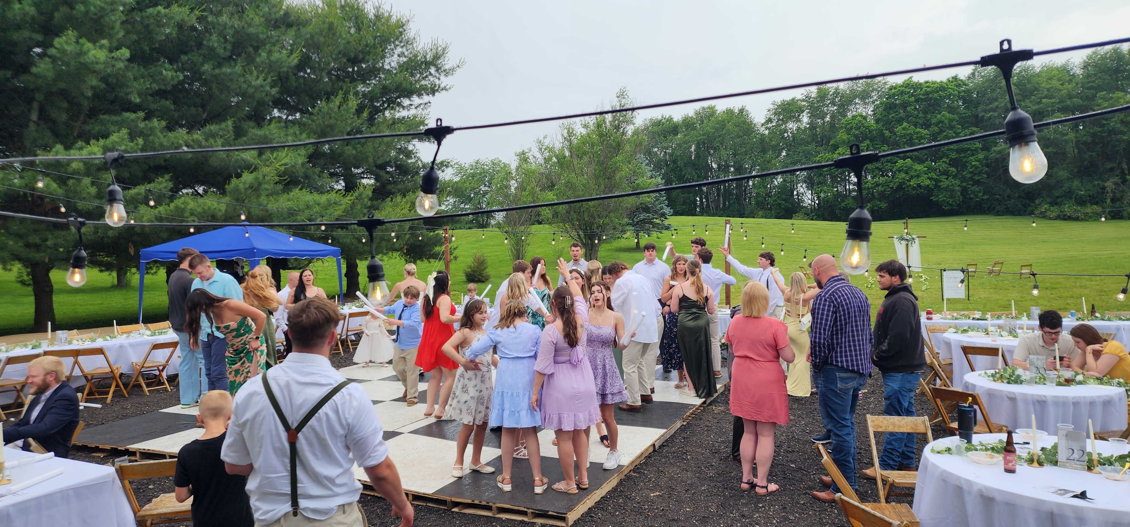 A diverse group of people dances on a wooden floor at an outdoor event, with tables set for dining and greenery in the background.