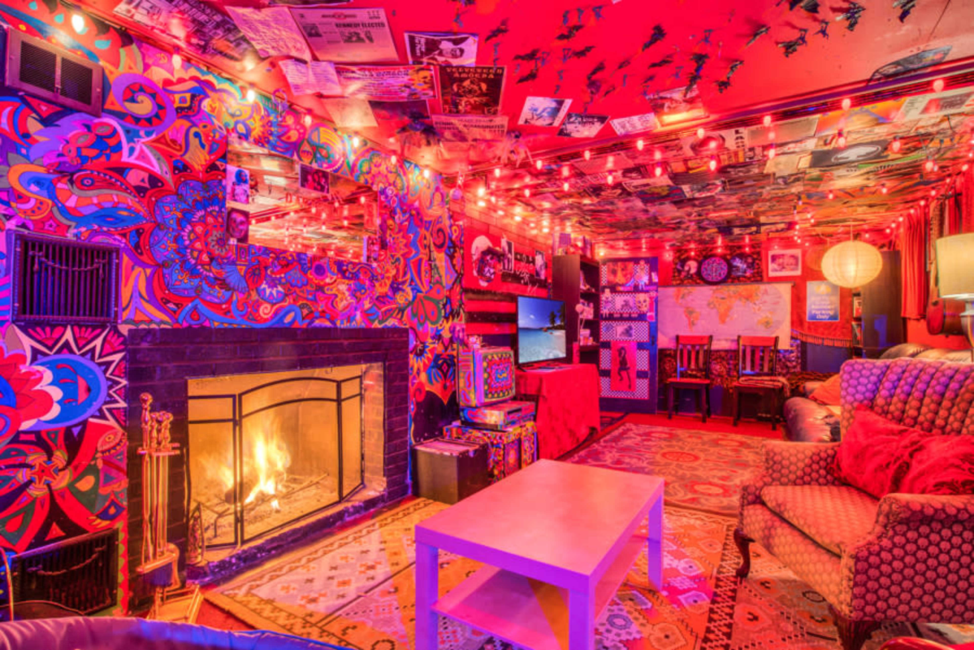 A brightly decorated living room features colorful artwork on the walls and ceiling, a fireplace with a glowing fire, and string lights illuminating the space.