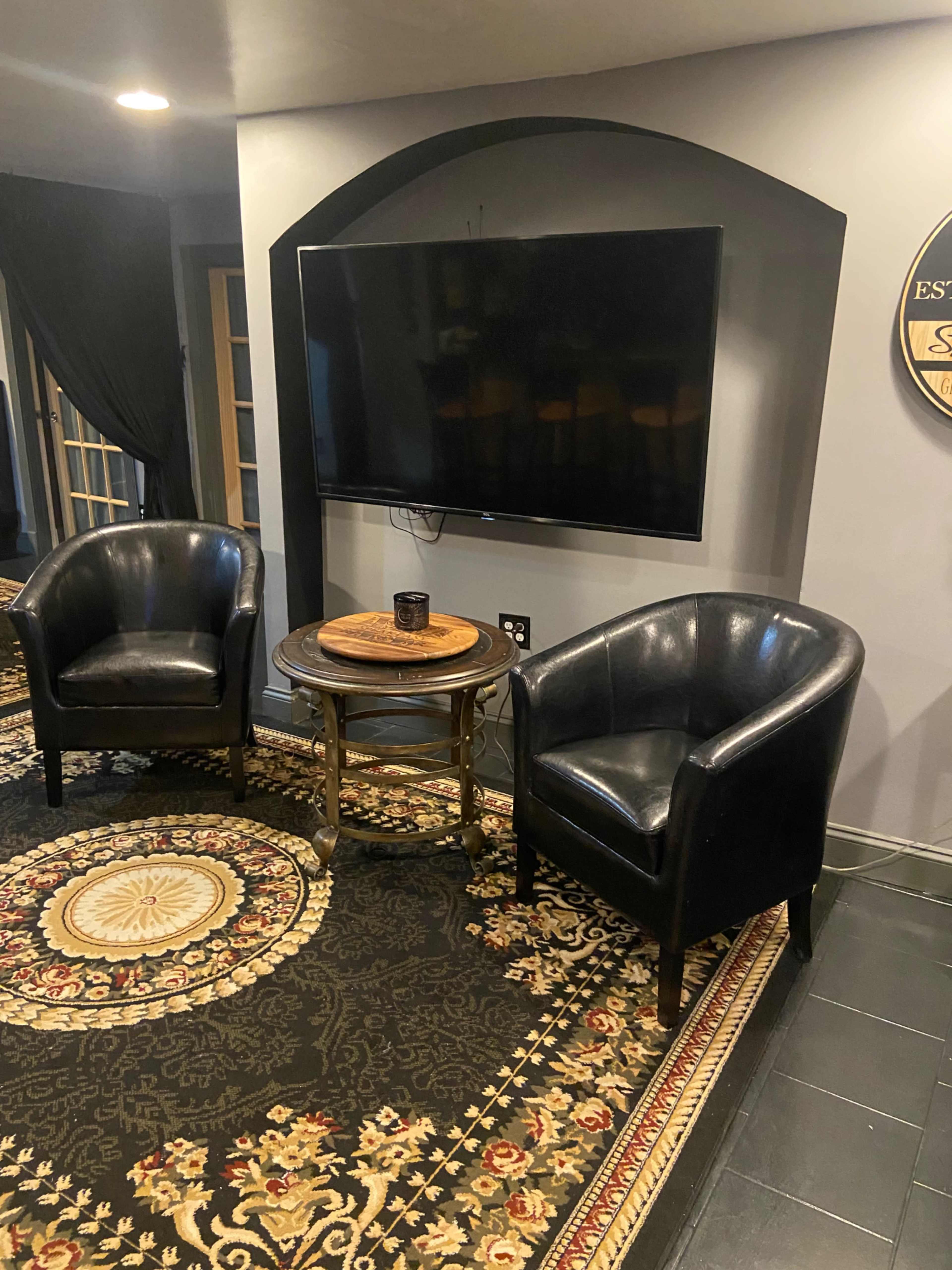 Two black leather armchairs facing a wall-mounted flat-screen TV, accompanied by a round wooden table on a patterned area rug.