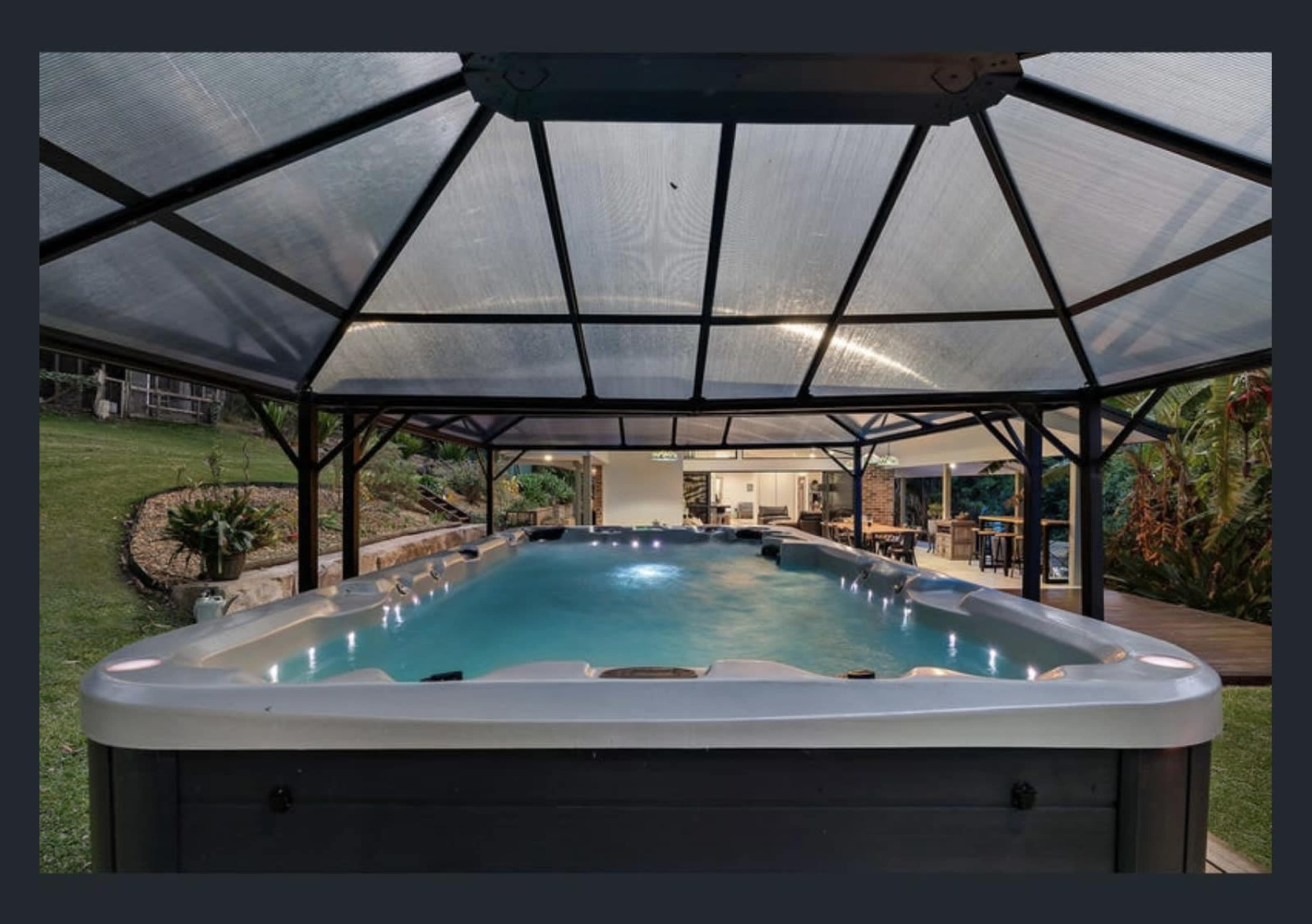 A spacious indoor swimming pool housed under a glass-roofed structure, surrounded by greenery and leading to an outdoor seating area.