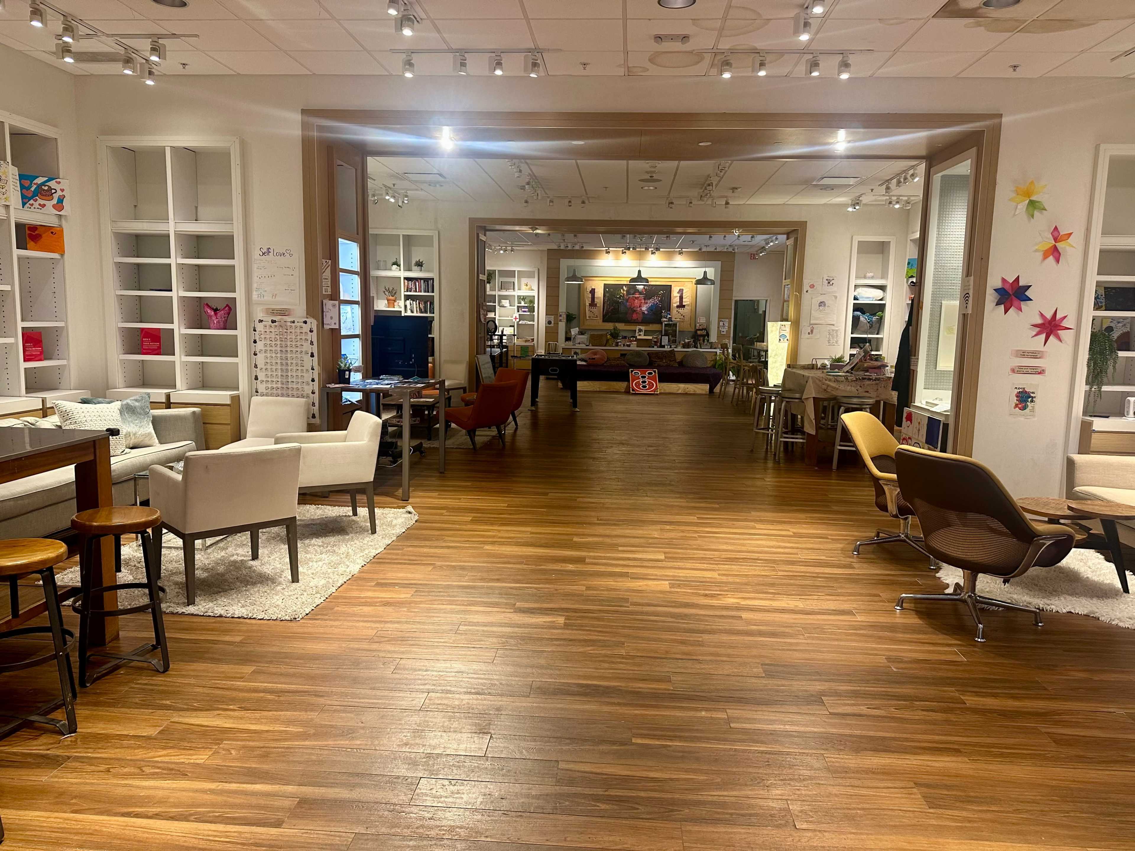 The image shows a spacious communal area with wooden flooring, featuring various seating arrangements, tables, and artwork on the walls.