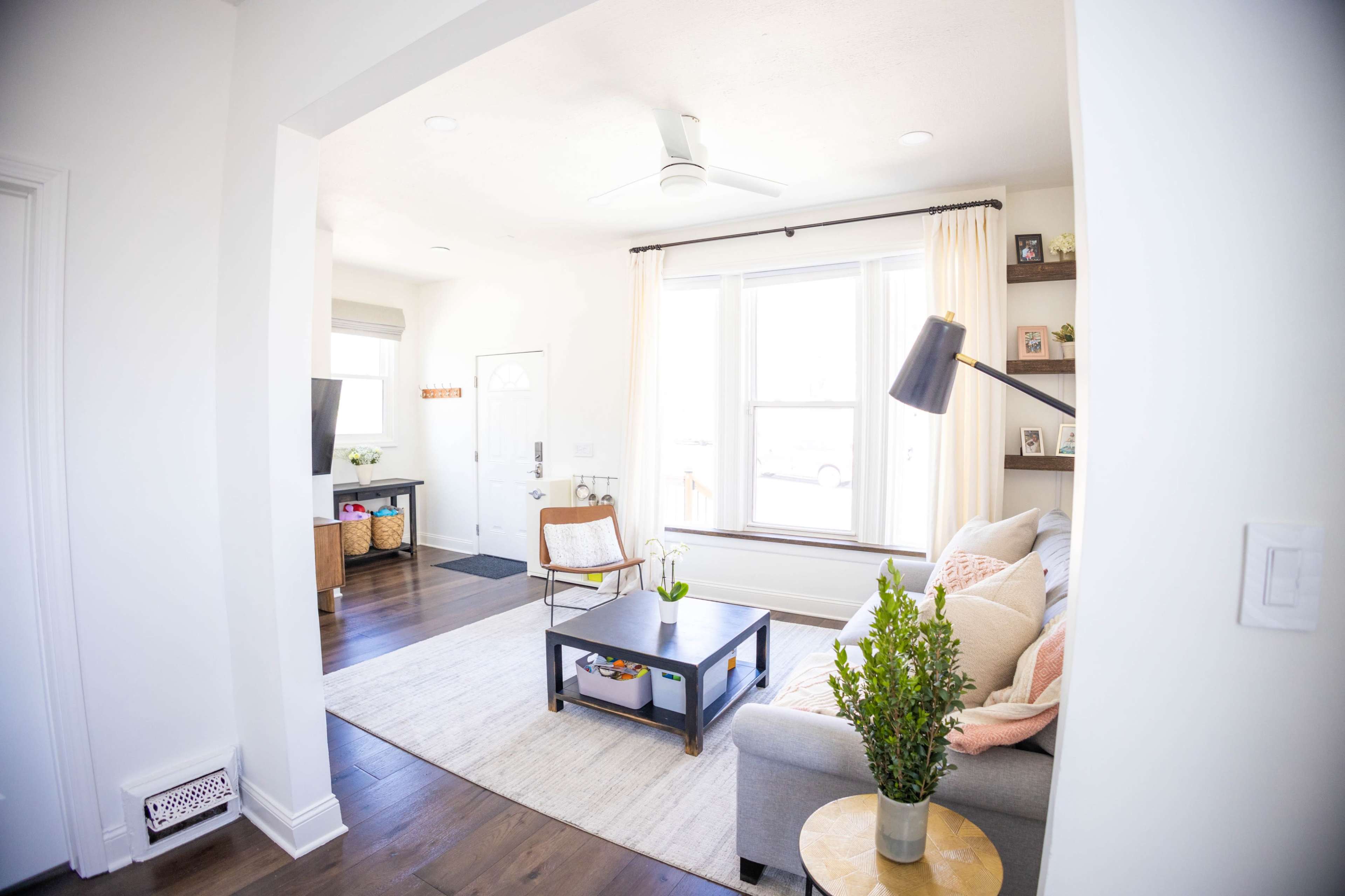 A bright living room features a gray sofa, a coffee table, and a large window with curtains, visible from an adjoining hallway.