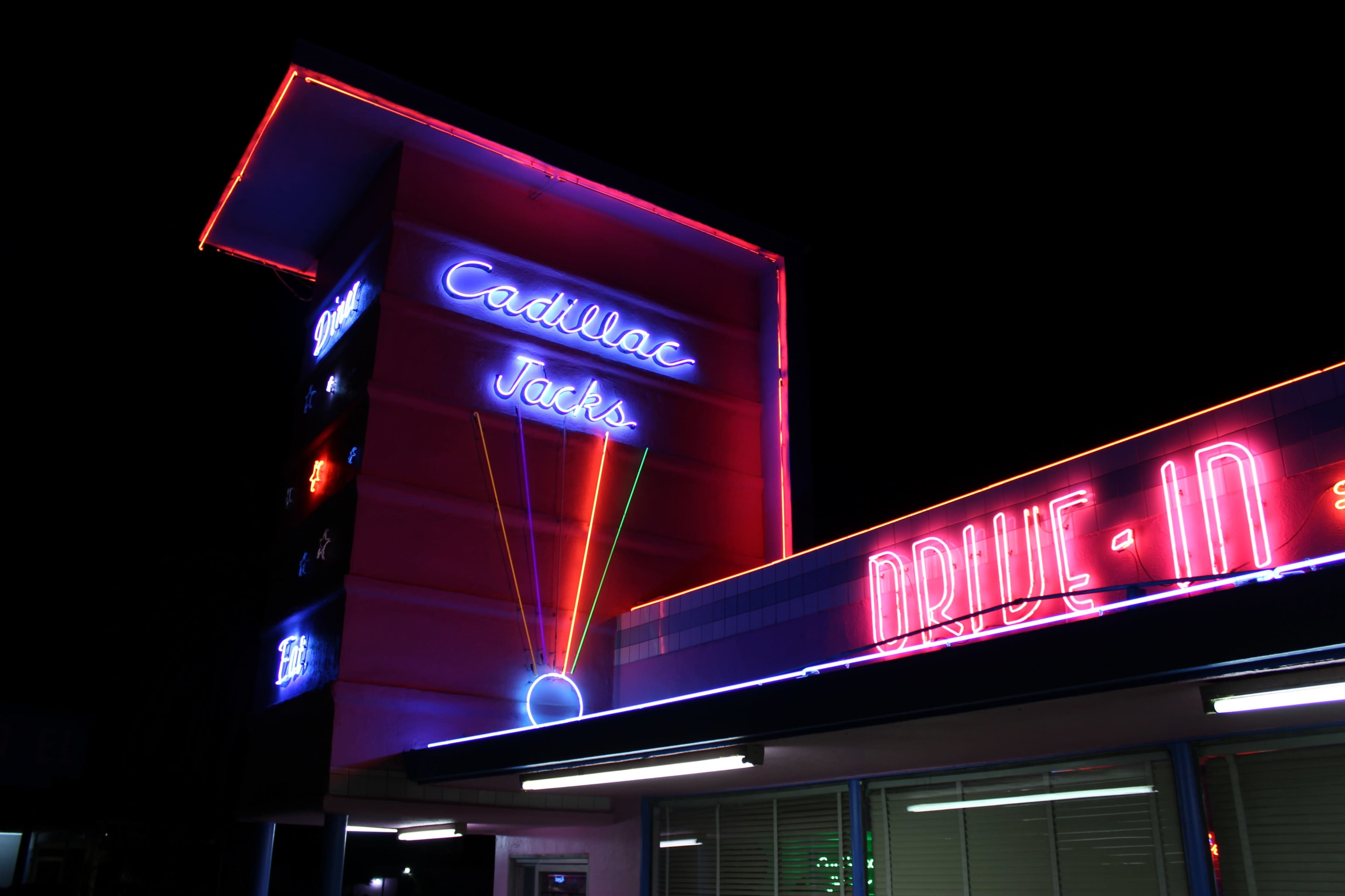 A brightly lit neon sign for "Cadillac Jacks" and "Drive-In" illuminates a building at night.