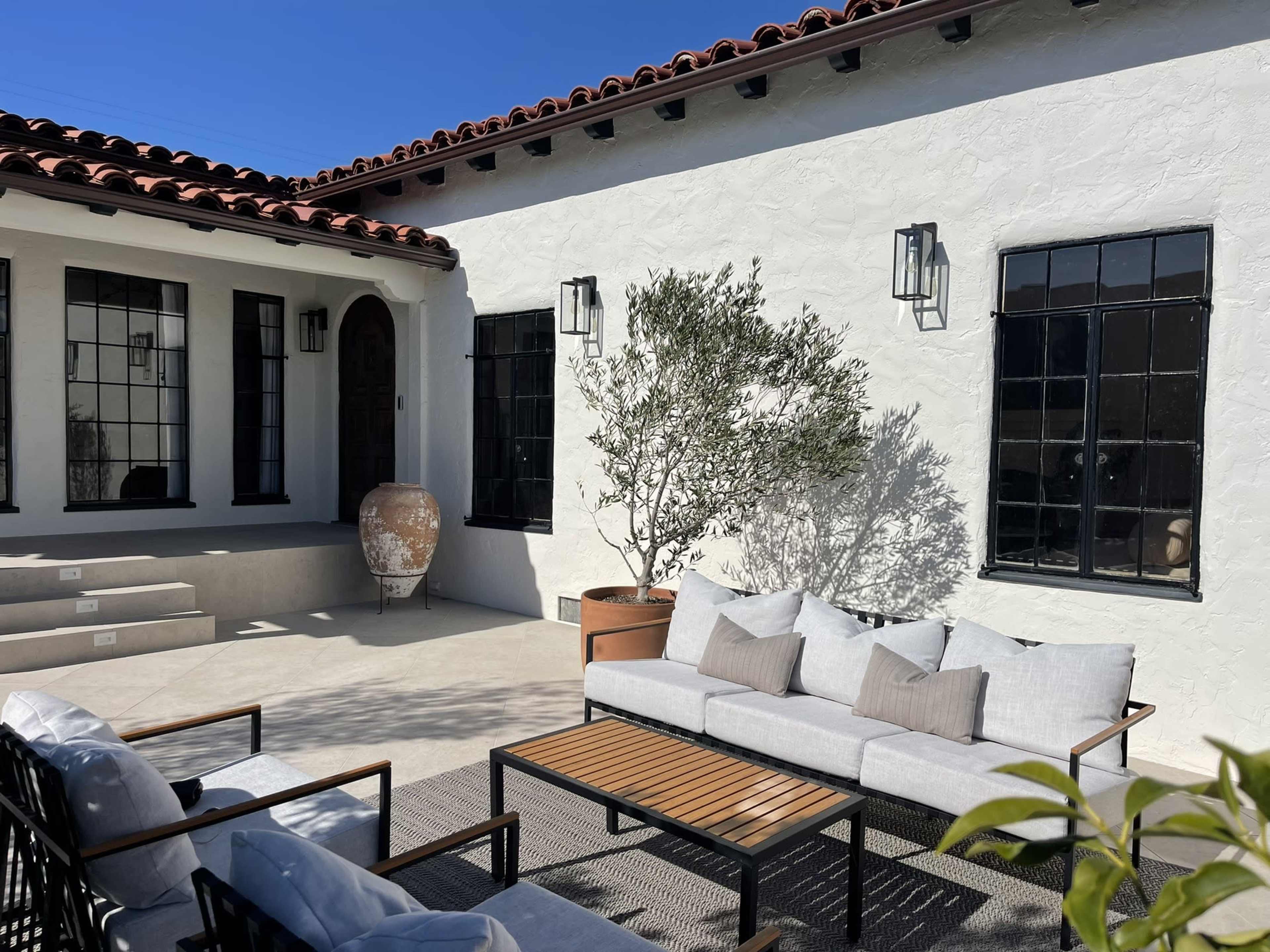 A patio area featuring modern outdoor furniture, a decorative pot, and a small tree beside a white stucco building with black window frames.