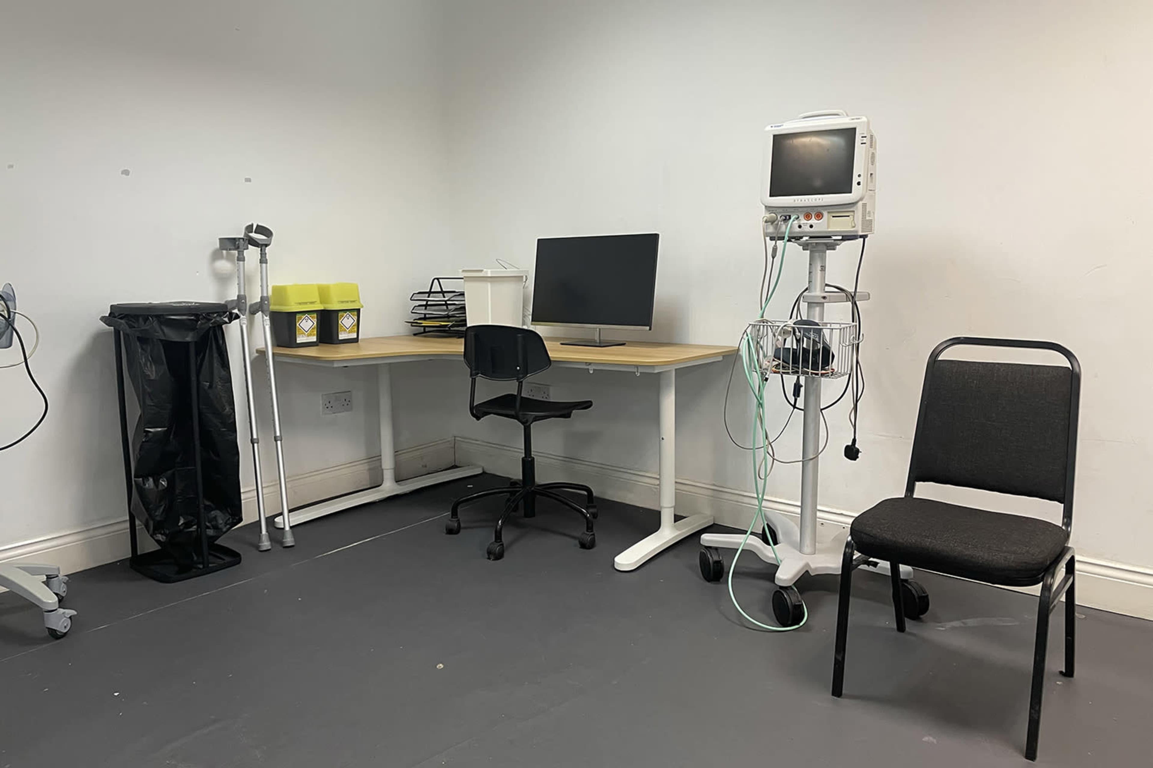 A clinical room with a desk and chair, a medical monitor, a trash can, and a black chair.