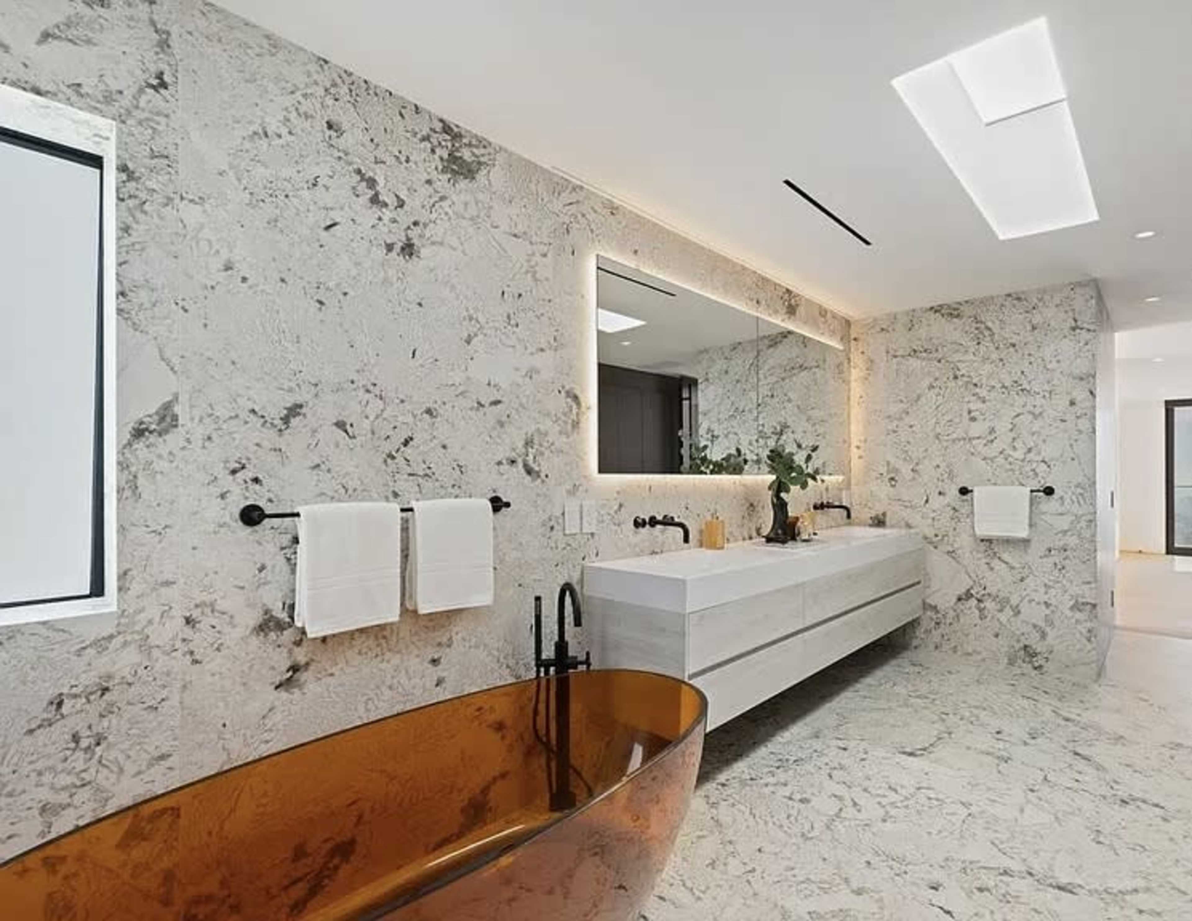 The image shows a modern bathroom featuring a freestanding amber-colored bathtub, a double vanity with a large mirror illuminated by soft lighting, and textured stone walls.