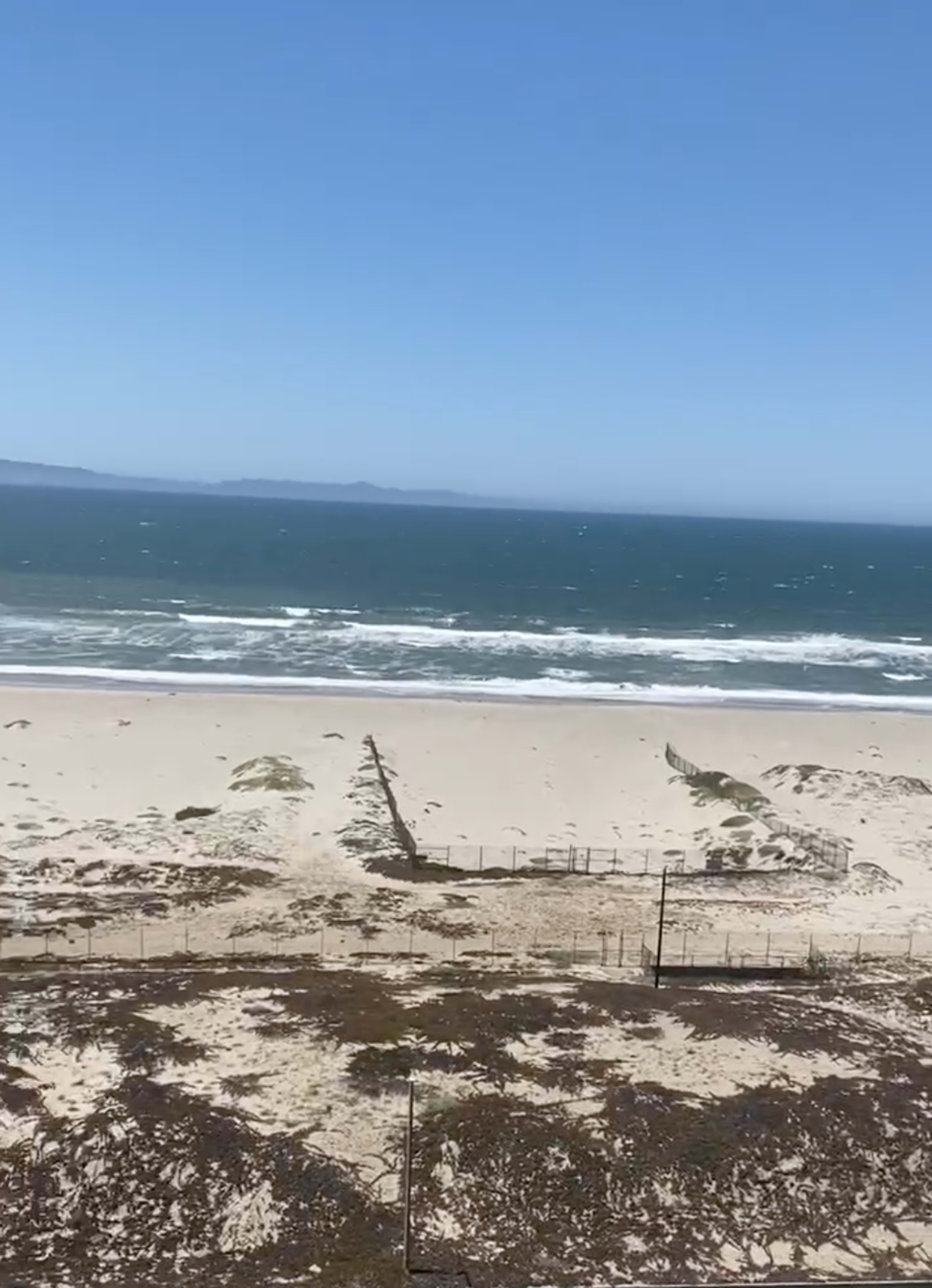 A sandy beach with gentle waves lapping at the shore under a clear blue sky, with distant mountains visible on the horizon.