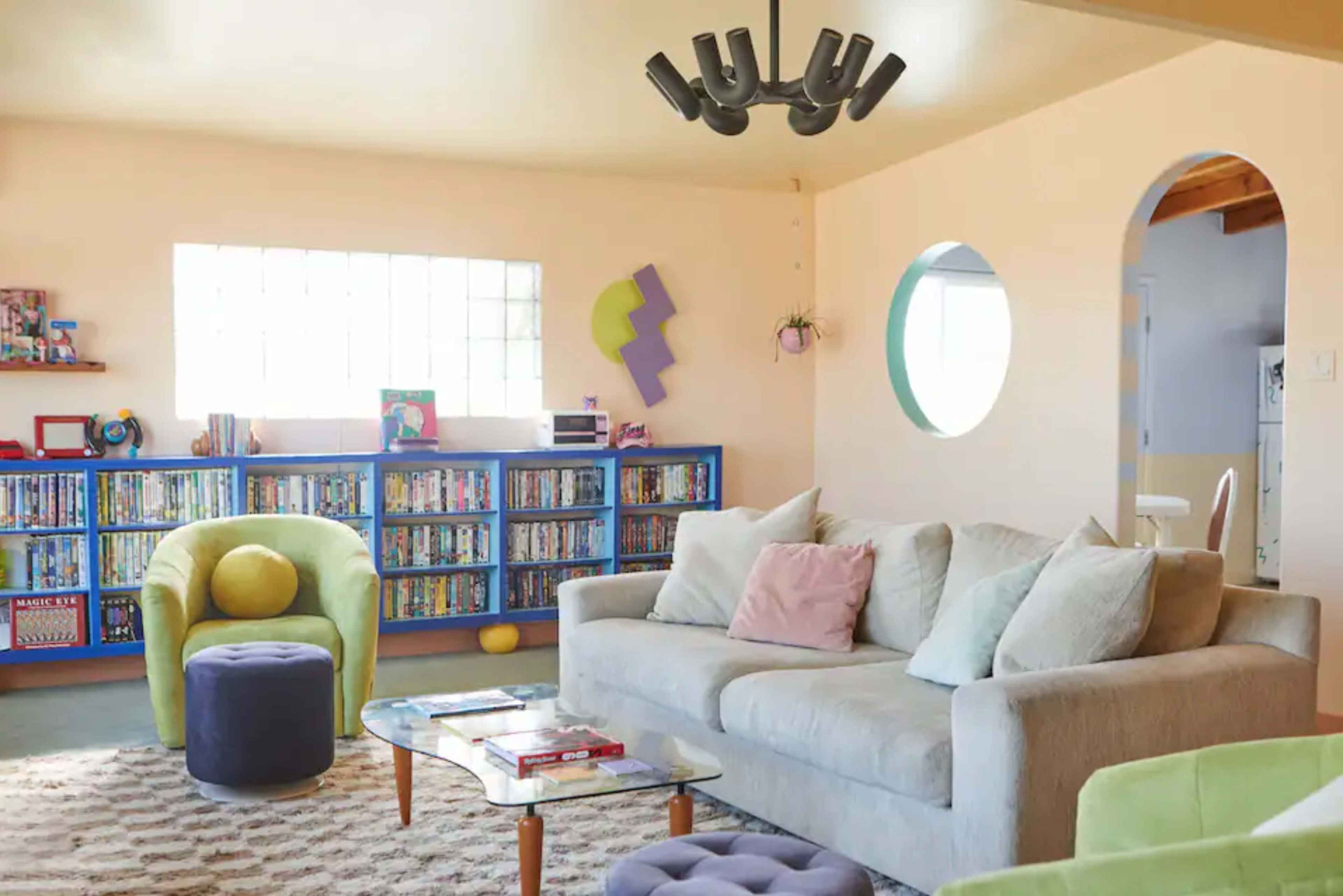 A bright living room features a light-colored sofa, colorful bookshelves filled with DVDs, and a round window, all anchored by a light fixture.