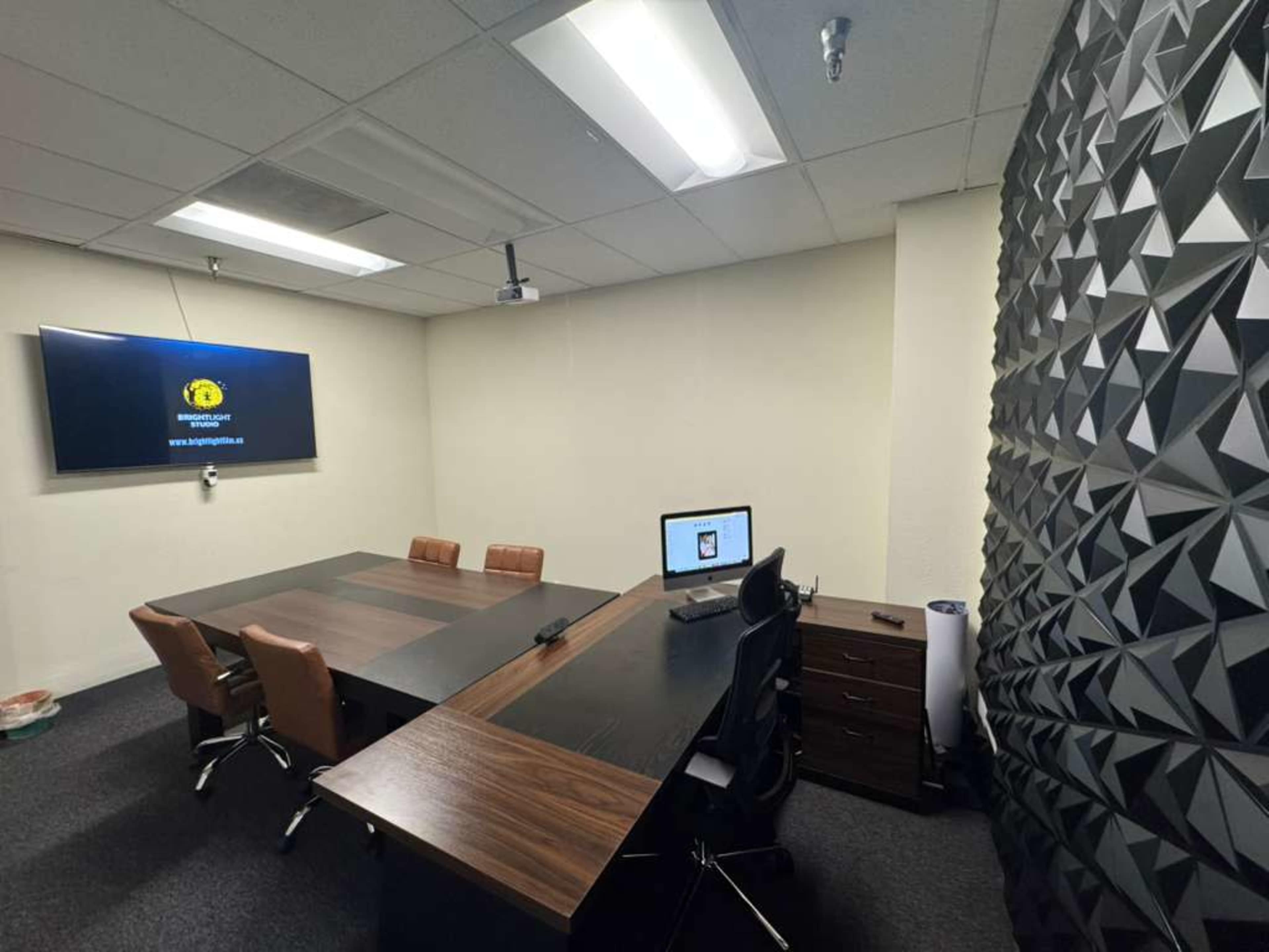 The image shows a modern conference room with a large table, chairs, a wall-mounted screen, and a textured feature wall.