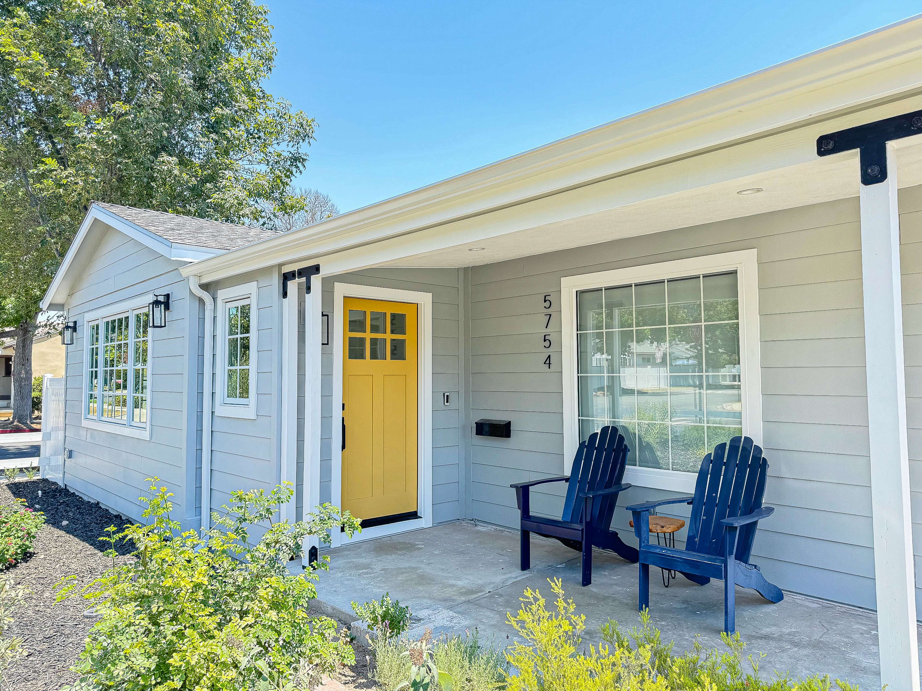 The image shows a gray house with a bright yellow front door and two blue Adirondack chairs on the porch.