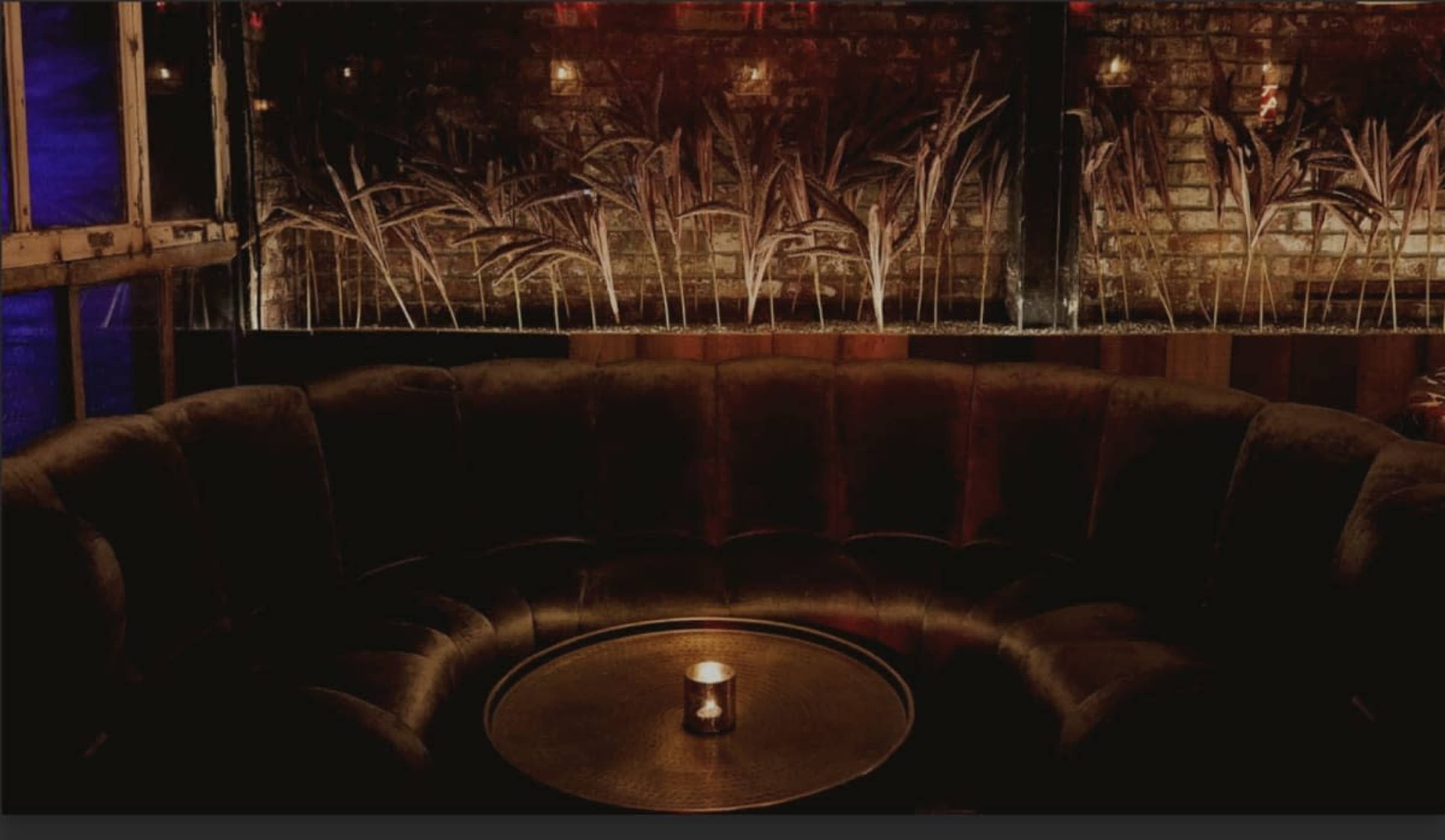 A circular, dark velvet seating area surrounds a small table with a lit candle, set against a backdrop of stylized wheat-like decor on a brick wall.
