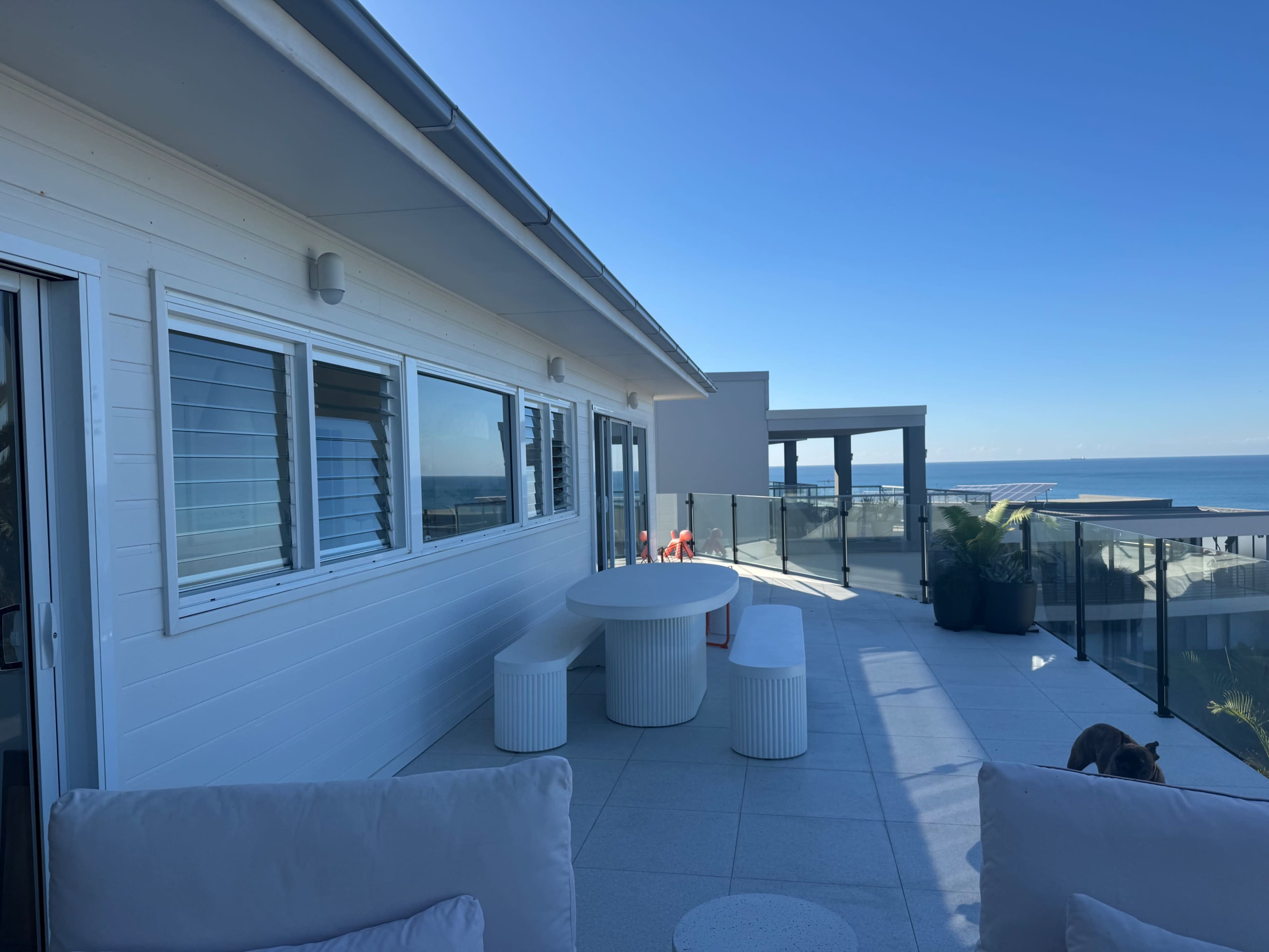 The image shows a terrace with a round white table and two cylindrical benches, overlooking a calm ocean under a clear blue sky.