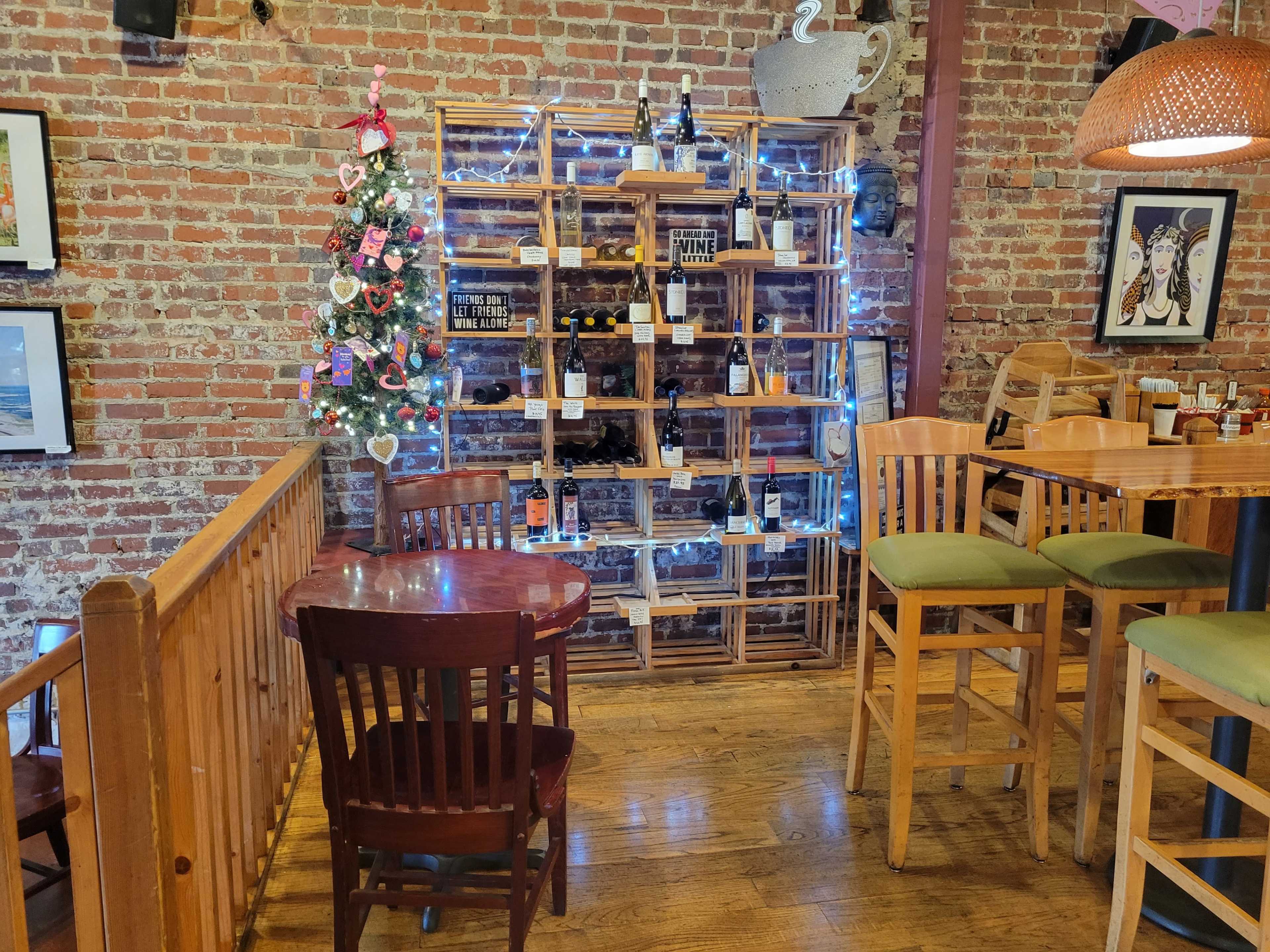 A wooden wine rack with bottles displayed is illuminated by string lights in a cozy restaurant setting, featuring a small table and chairs nearby.