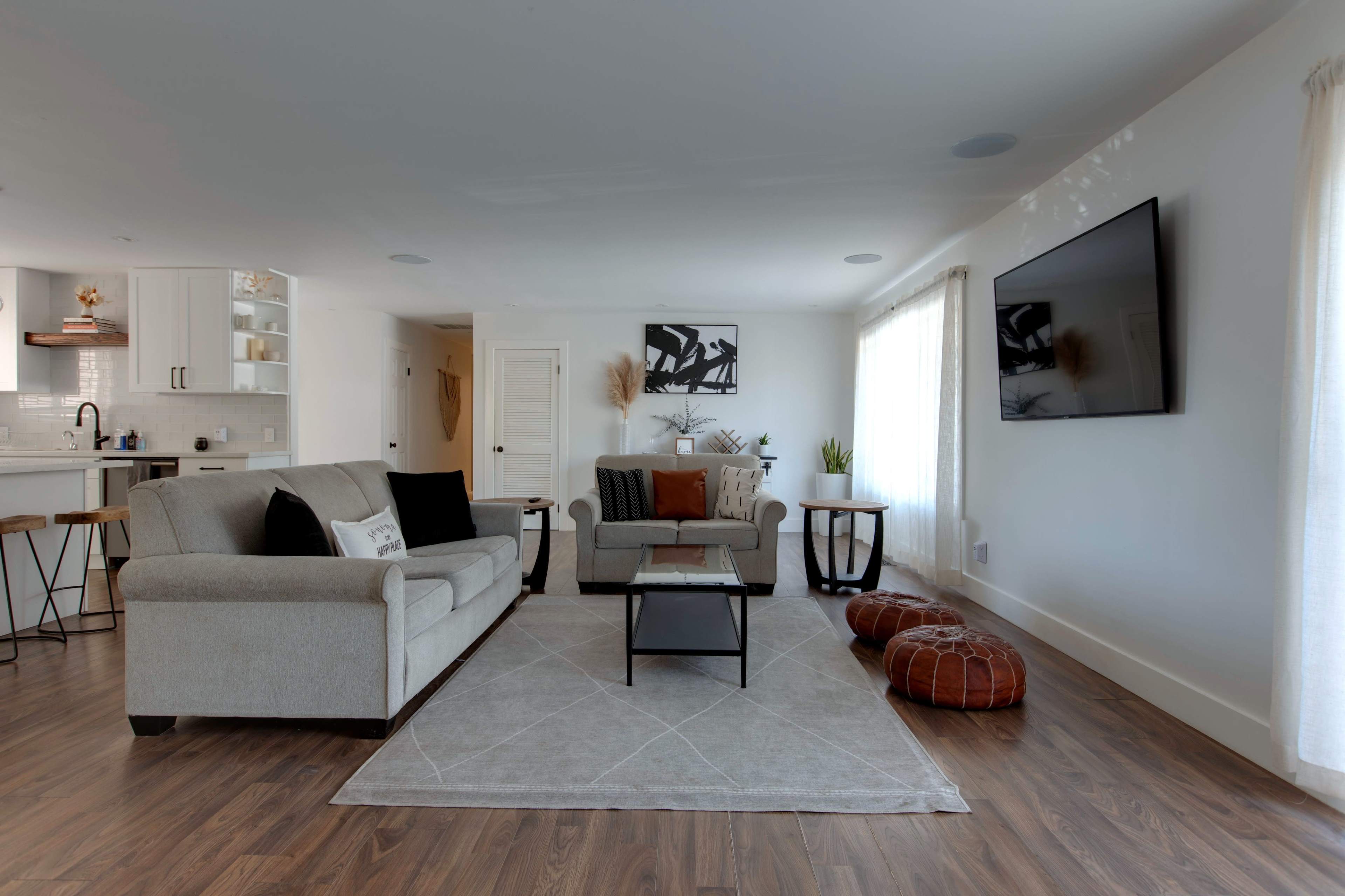 The living room features a sofa, a coffee table, two poufs, and a large television mounted on the wall, with natural light coming through the windows.