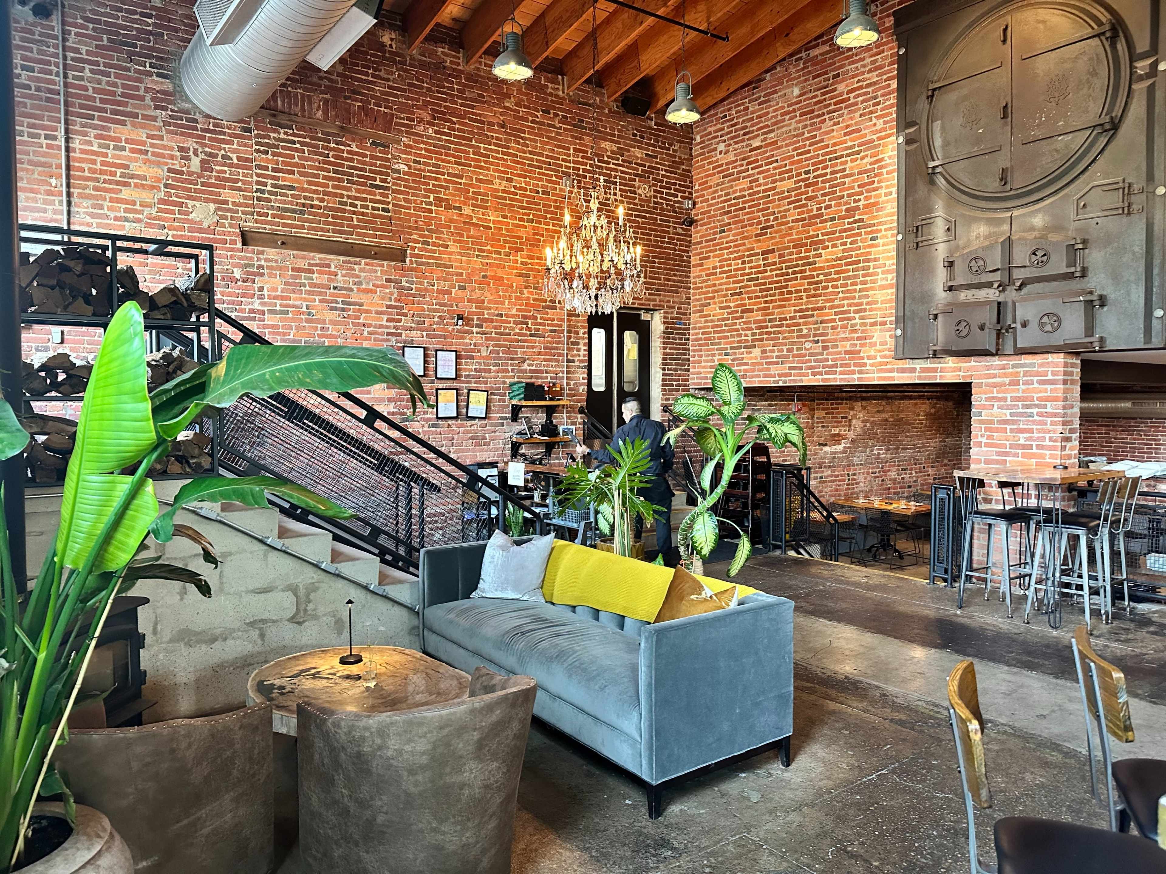 The image shows a stylish café interior with exposed brick walls, a chandelier, and modern furnishings, including a gray sofa and potted plants.