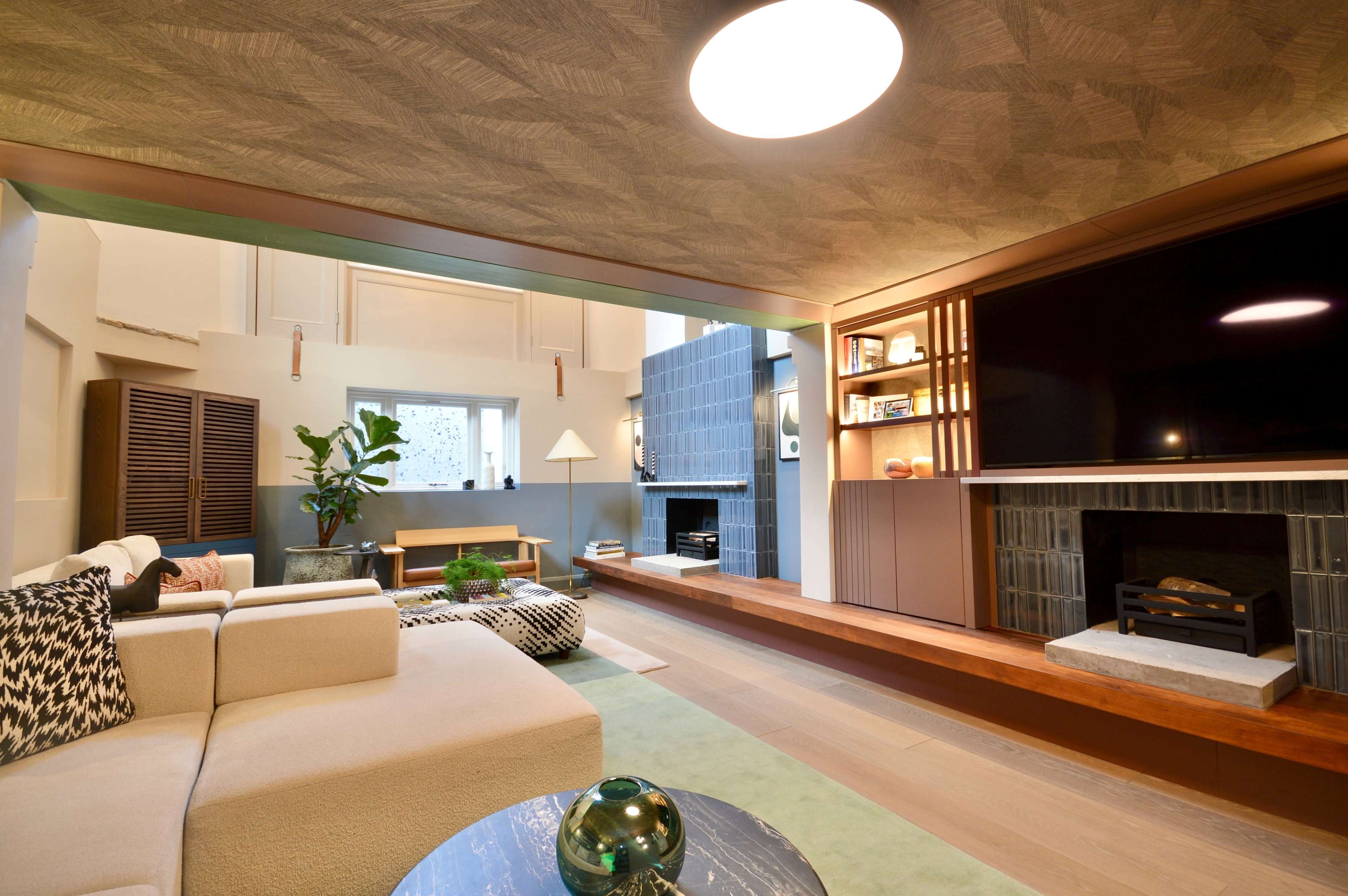 The image shows a modern living room with a beige sofa, a large television mounted on a wooden shelf, and a fireplace beneath a stylish blue tiled wall.