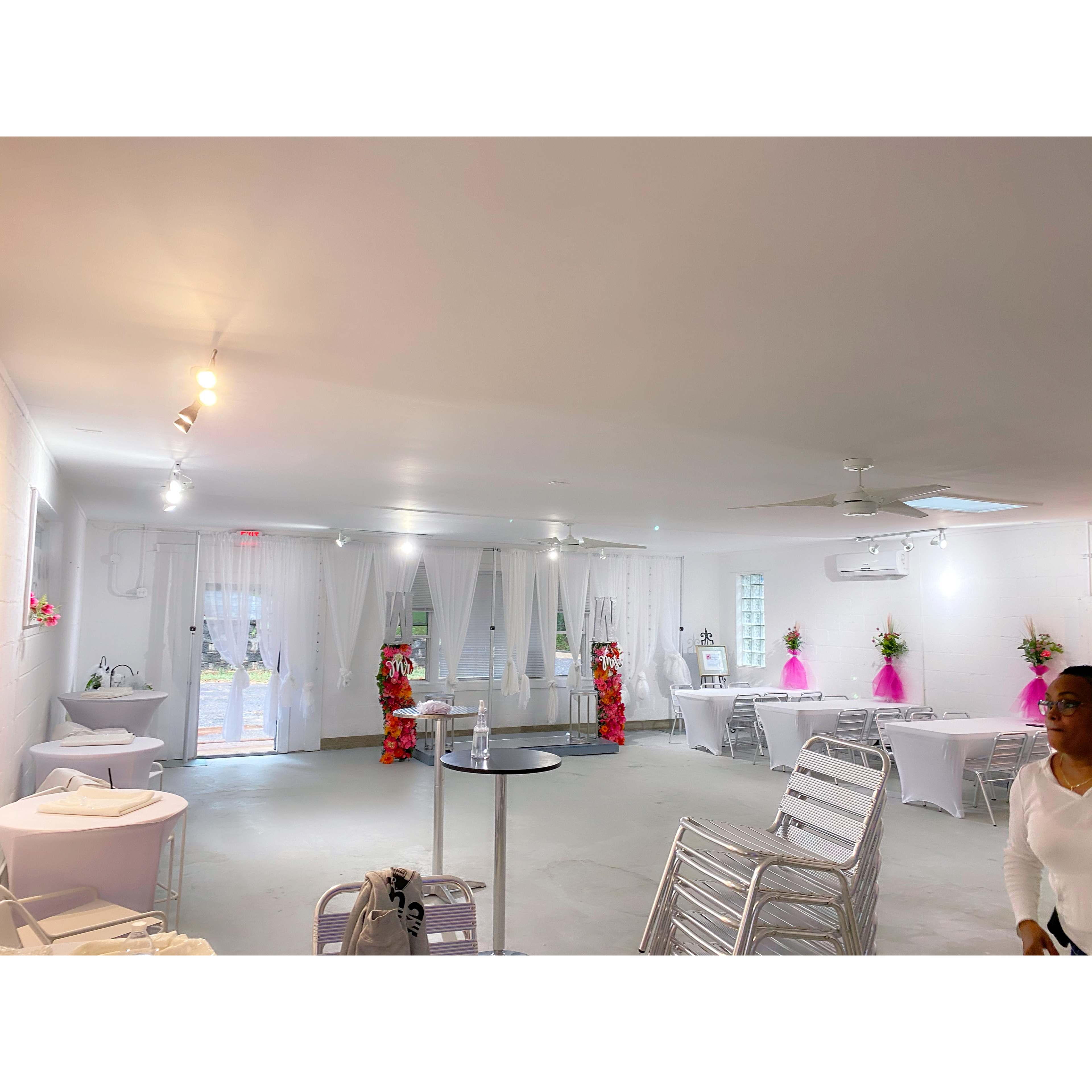 The image shows an indoor event space with white walls, several tables covered in white cloths, and decorative floral arrangements at either end.