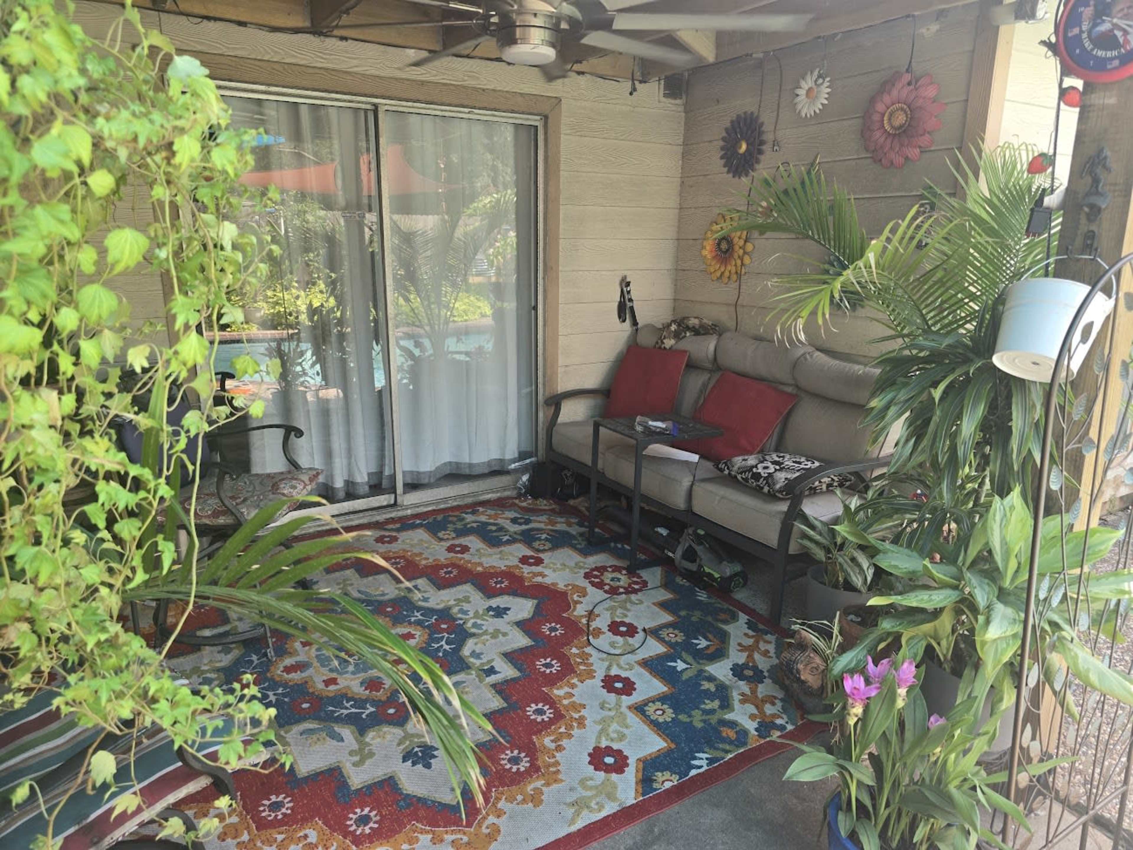 The image shows a cozy porch area with a sofa, decorative pillows, a vibrant rug, and potted plants.