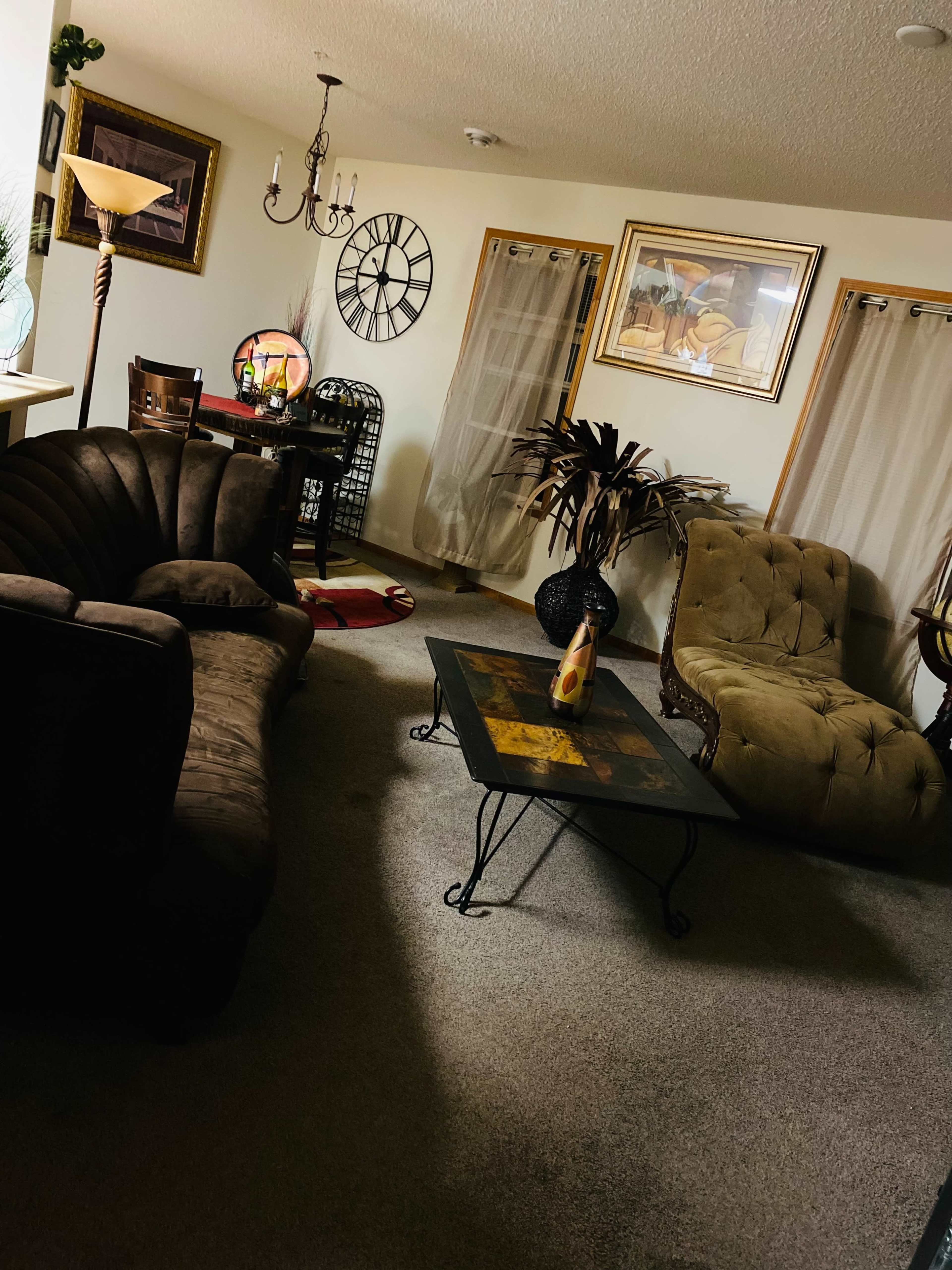 A living room with a brown sofa, an armchair, a glass coffee table, a dining area with a clock on the wall, and decor items including a lamp and plants.