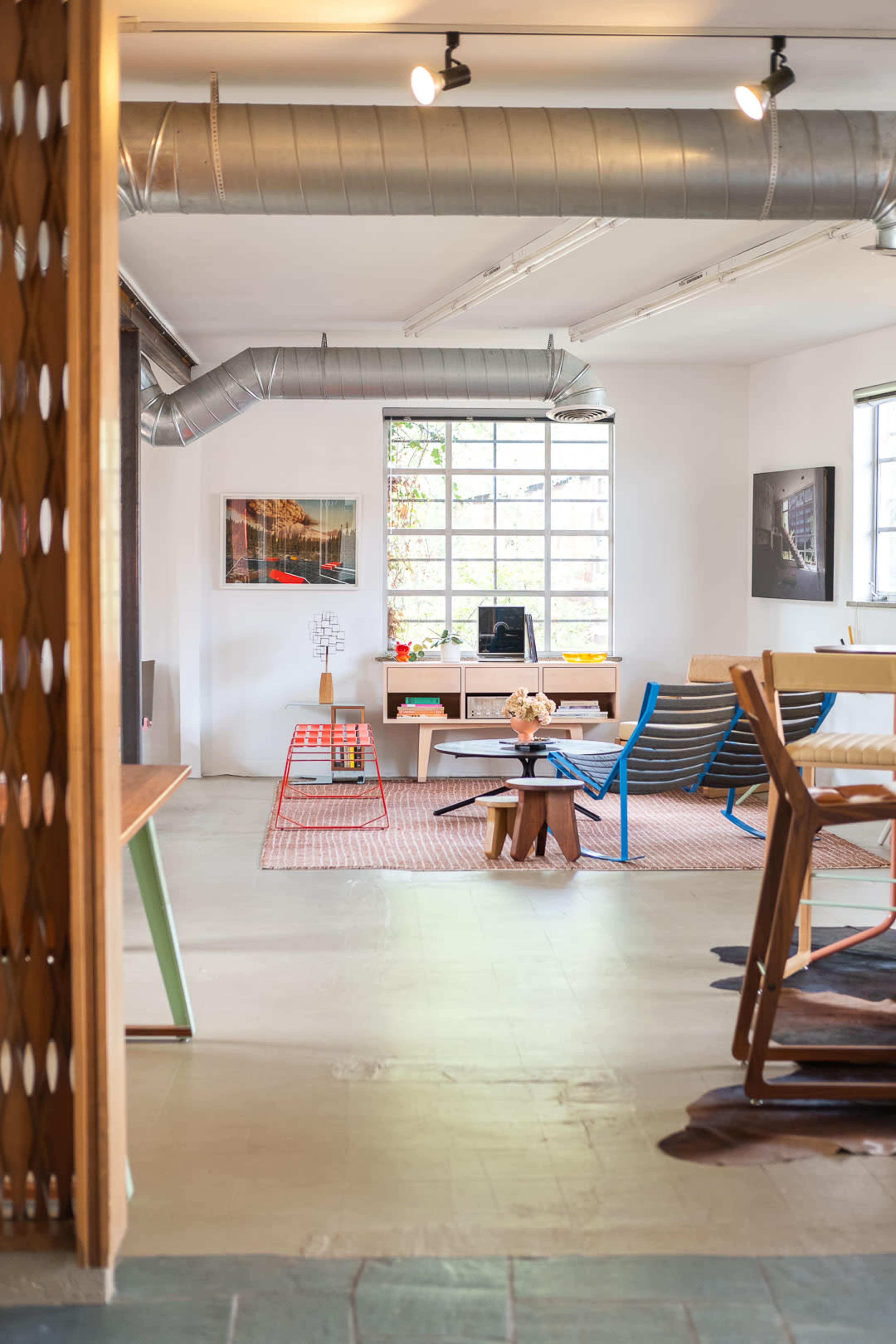 The image shows a modern office interior with a variety of furniture, including desks, chairs, and a coffee table, all arranged around a central area that features large windows and industrial-style ductwork.