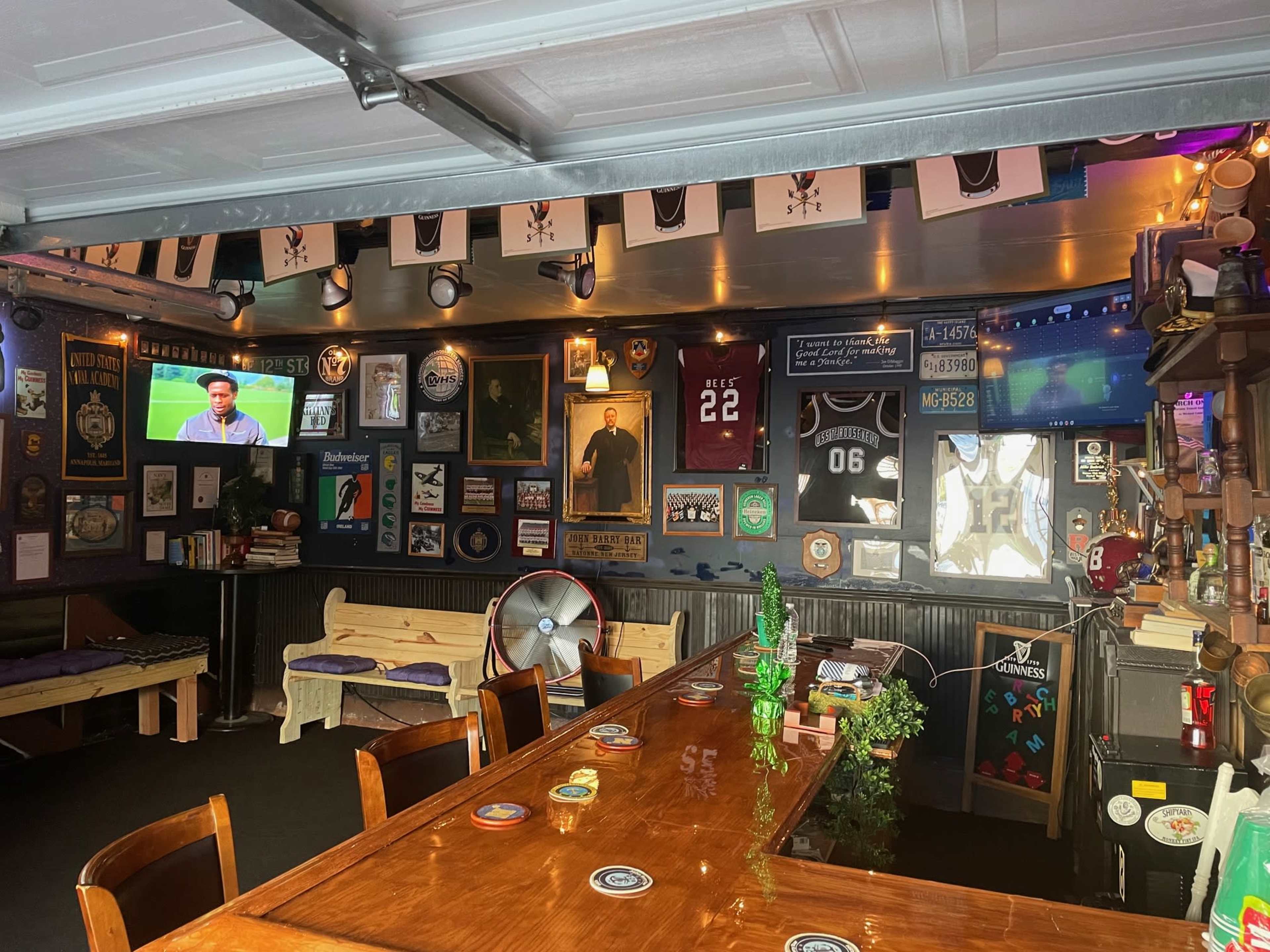 The image depicts a decorated garage with a wooden bar, various sports jerseys and memorabilia on the walls, and a television displaying a program.