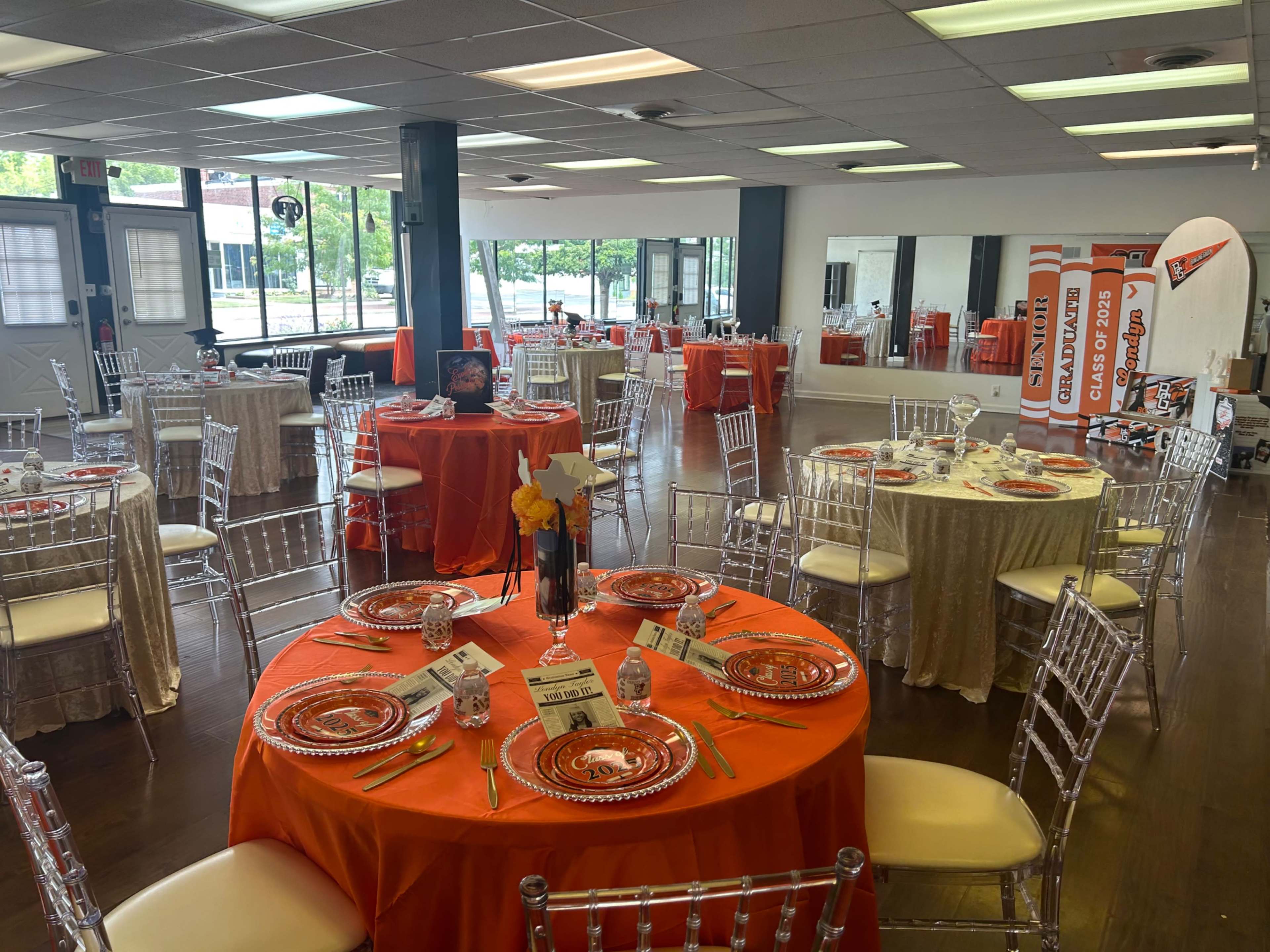 The image shows an event space set up with several circular tables adorned with red and beige tablecloths, each decorated with place settings and floral centerpieces.