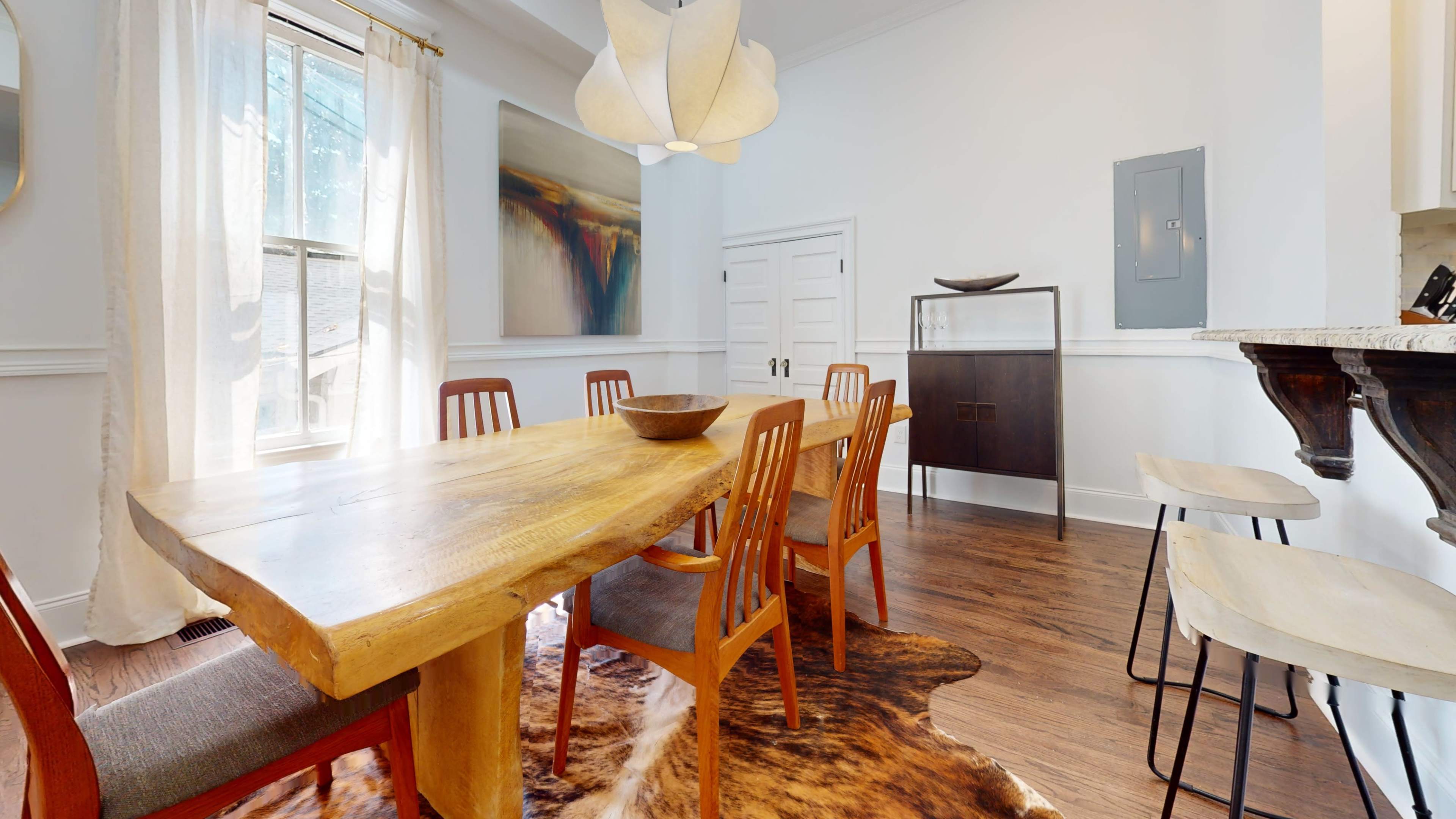 A wooden dining table with eight chairs is positioned in a brightly lit room featuring a large window and minimalist decor.