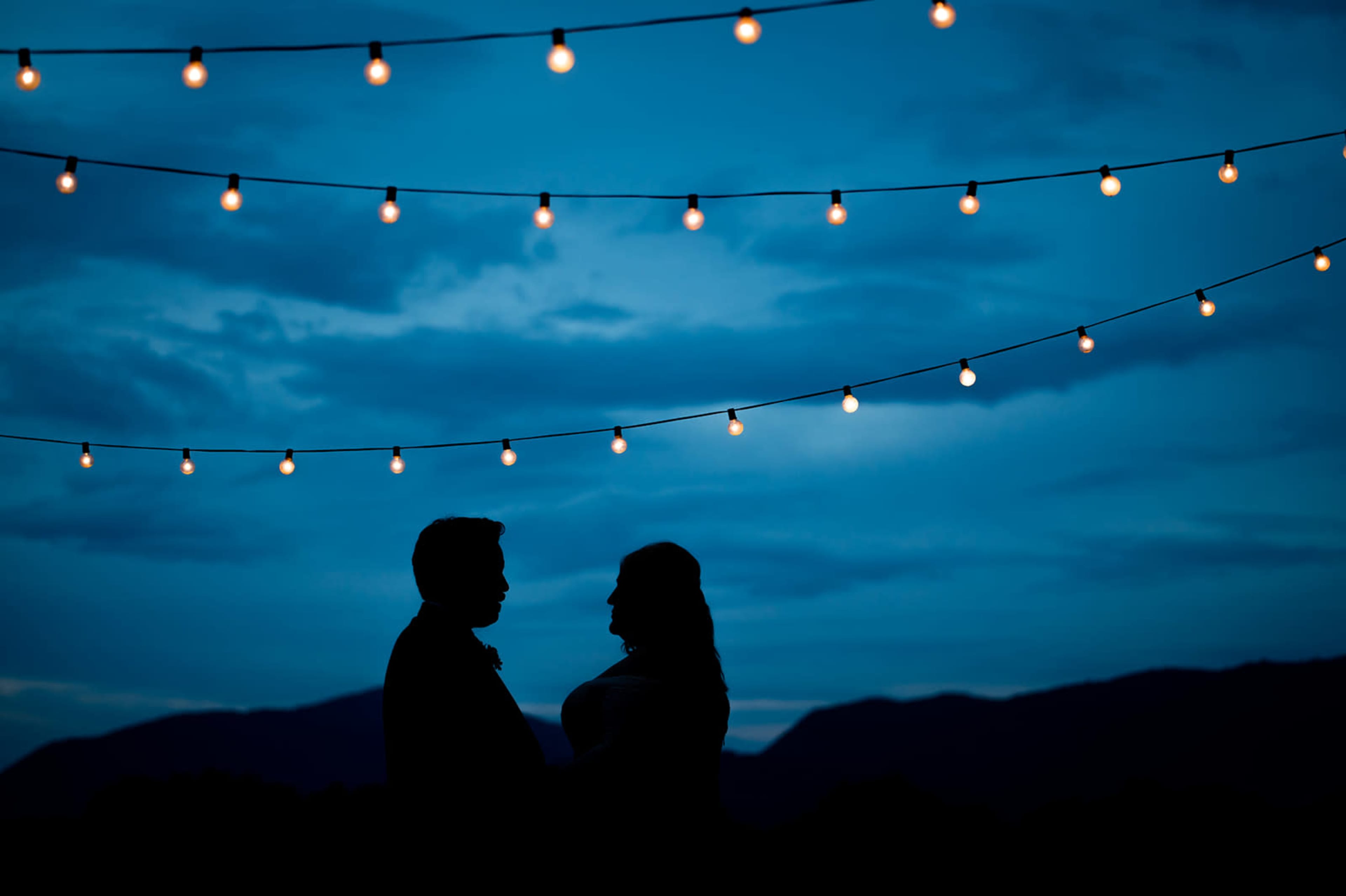 A couple stands in silhouette under string lights against a twilight sky with mountains in the background.