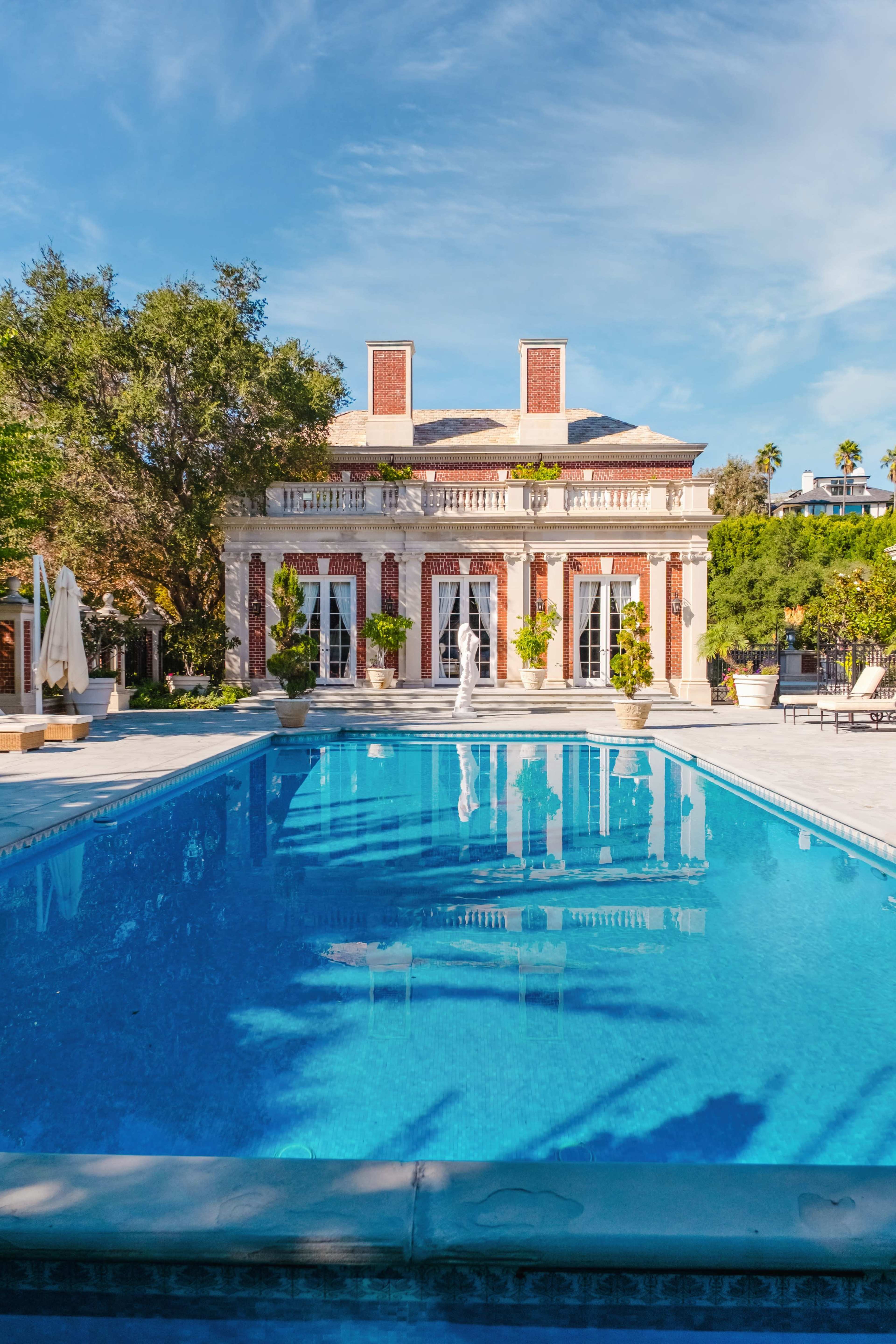 The image shows a luxurious swimming pool in front of an elegant, two-story mansion surrounded by lush greenery and palm trees.