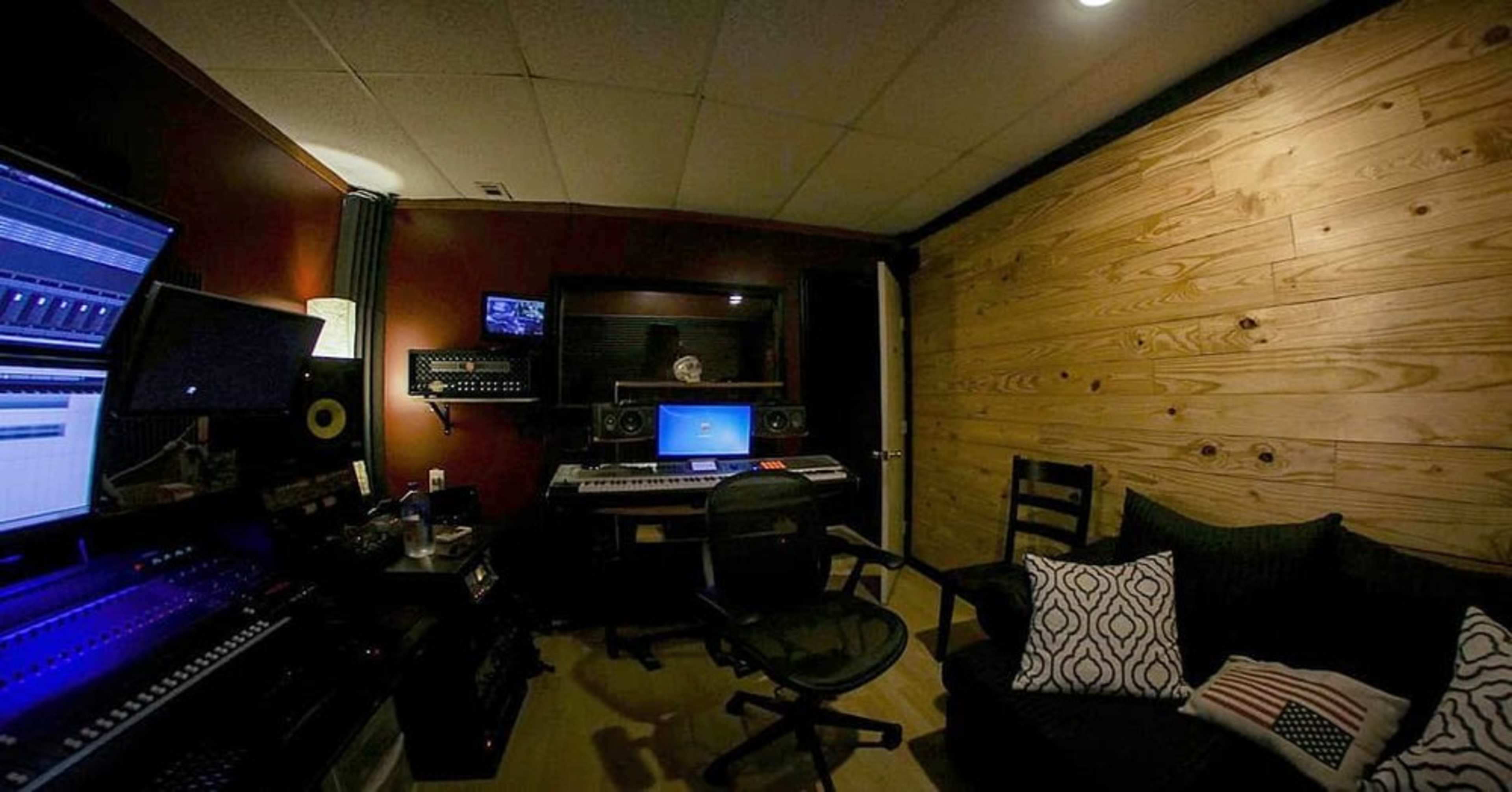 The image shows a music production studio featuring a computer desk with audio equipment, monitors, and a cozy seating area with a couch and decorative pillows.