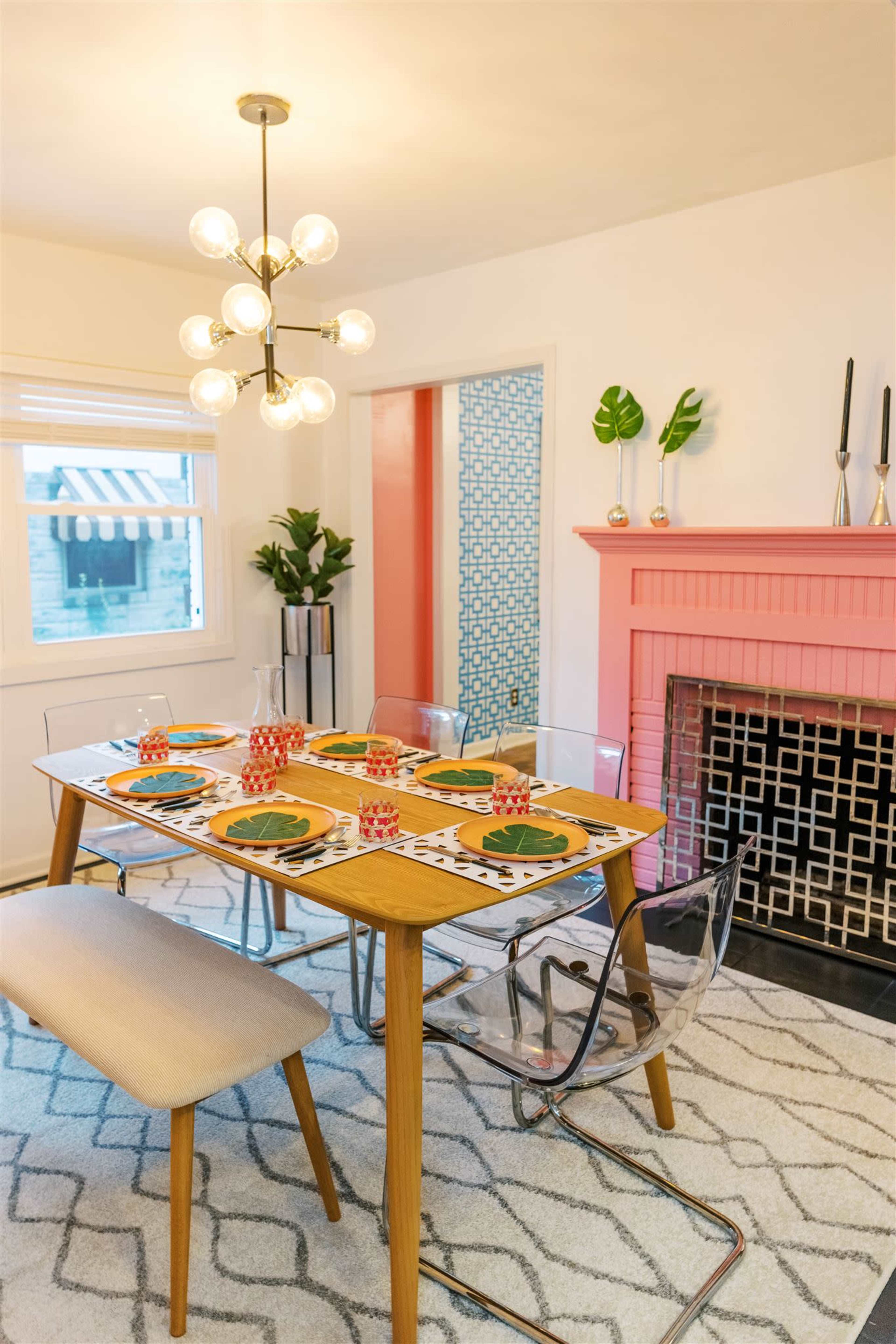 A modern dining room features a wooden table set with colorful plates and a pink accent wall with a fireplace.