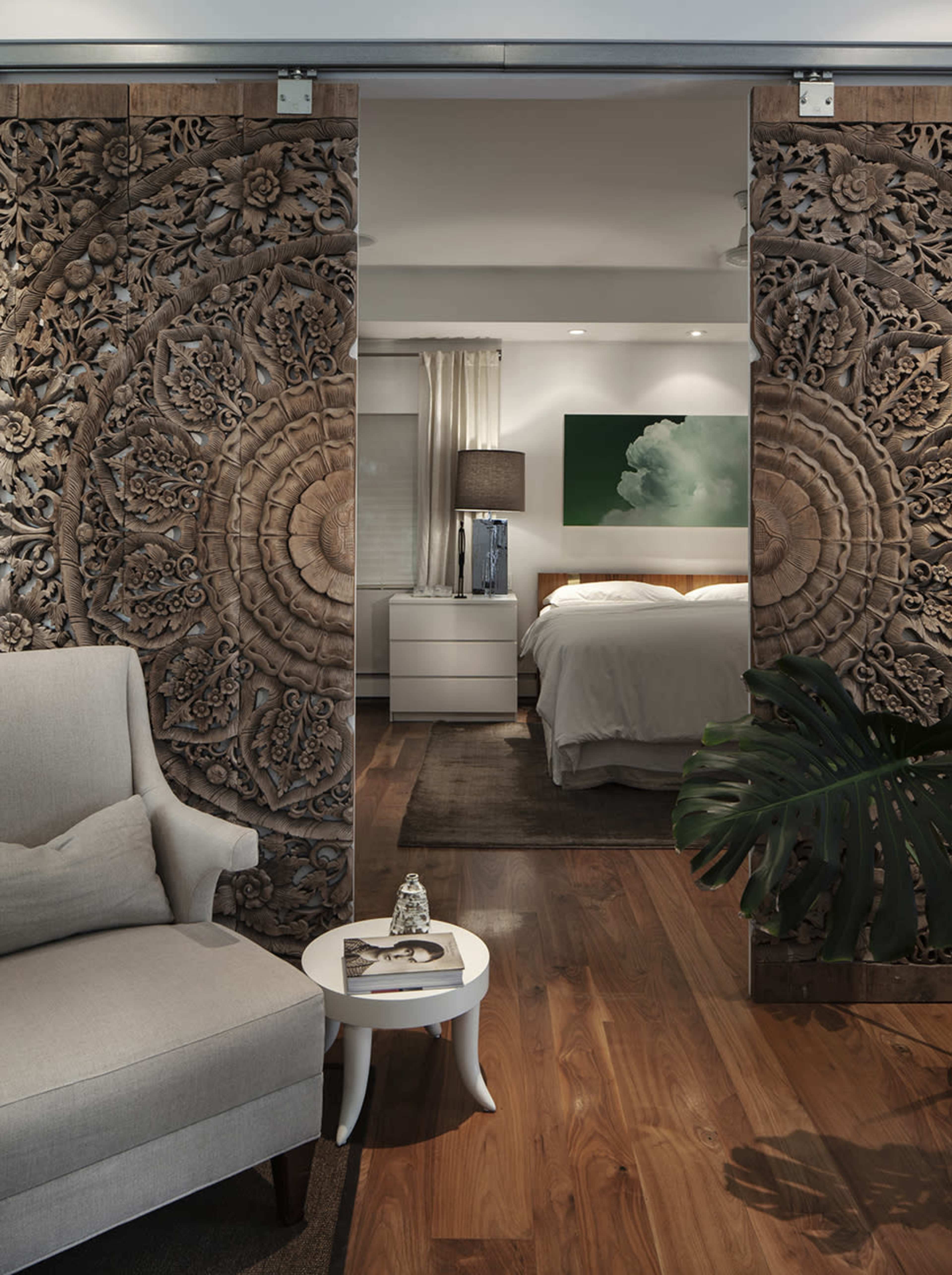 The image shows a room with intricately carved wooden panels separating a living area from a bedroom, featuring a light-colored armchair and a small white table.