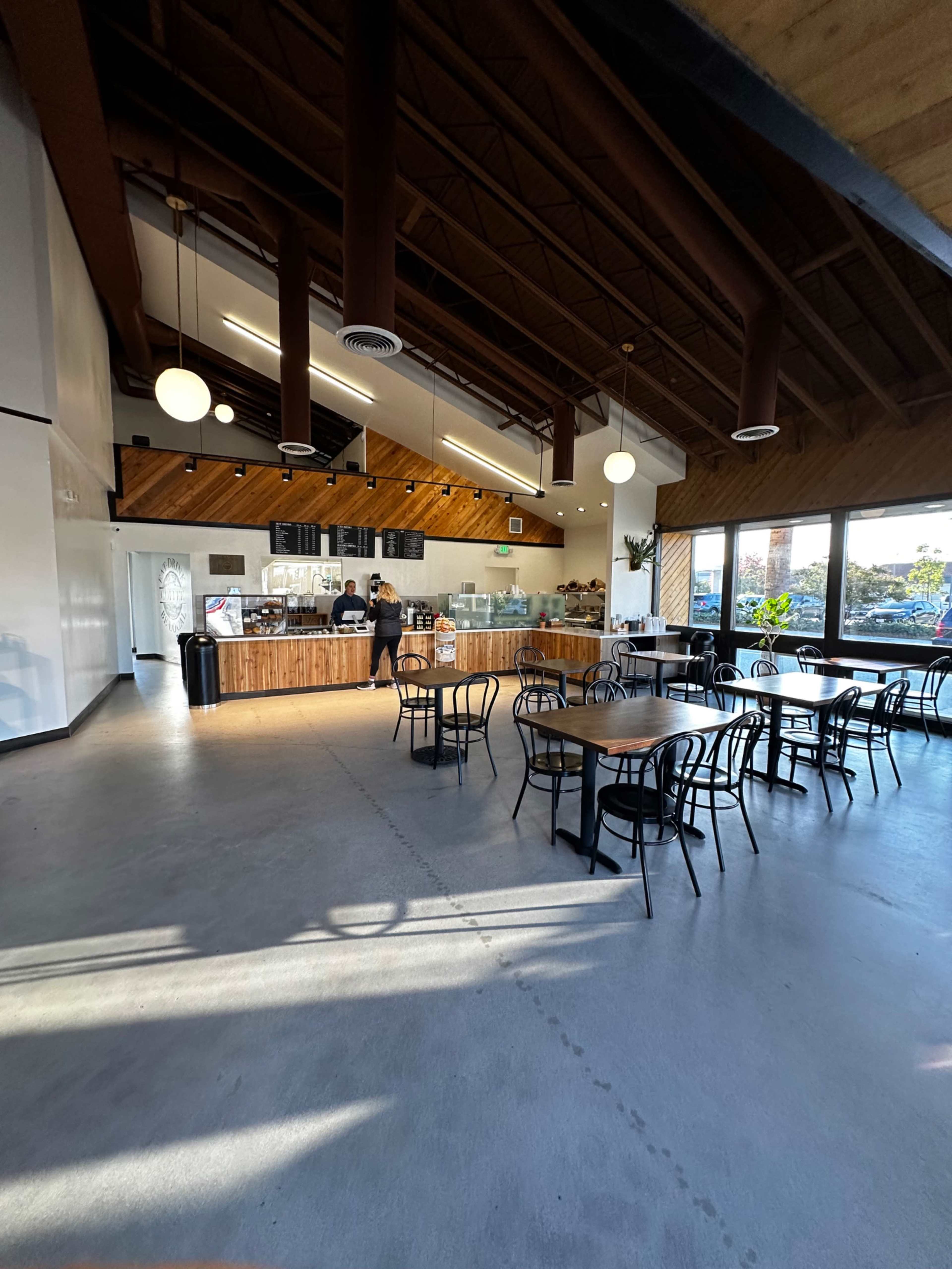 A spacious cafe features wooden accents, multiple tables, and a service counter where a staff member is preparing orders.