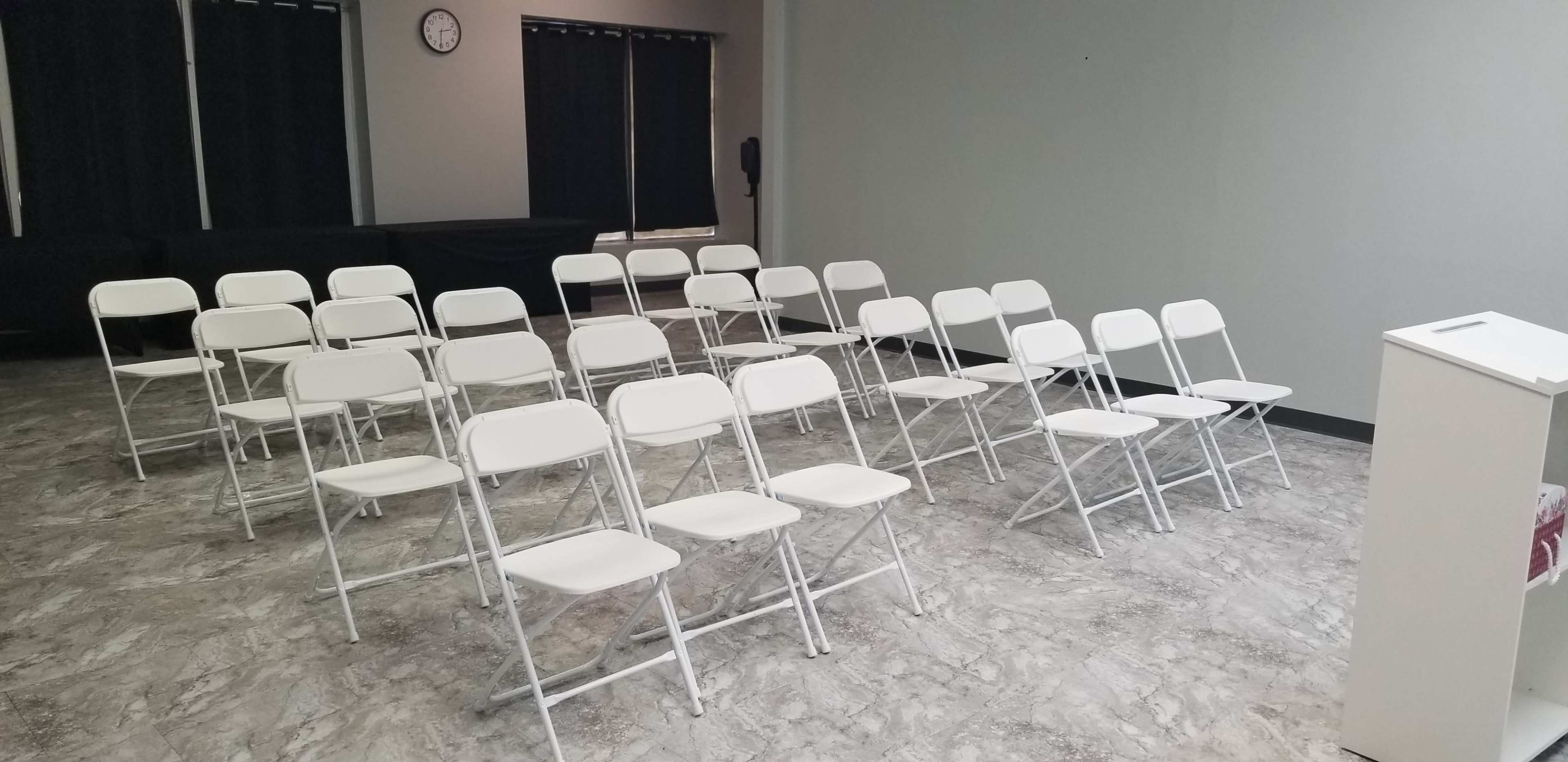 The image shows a room with white folding chairs arranged in rows facing a small podium, with a clock on the wall and dark curtains covering the windows.