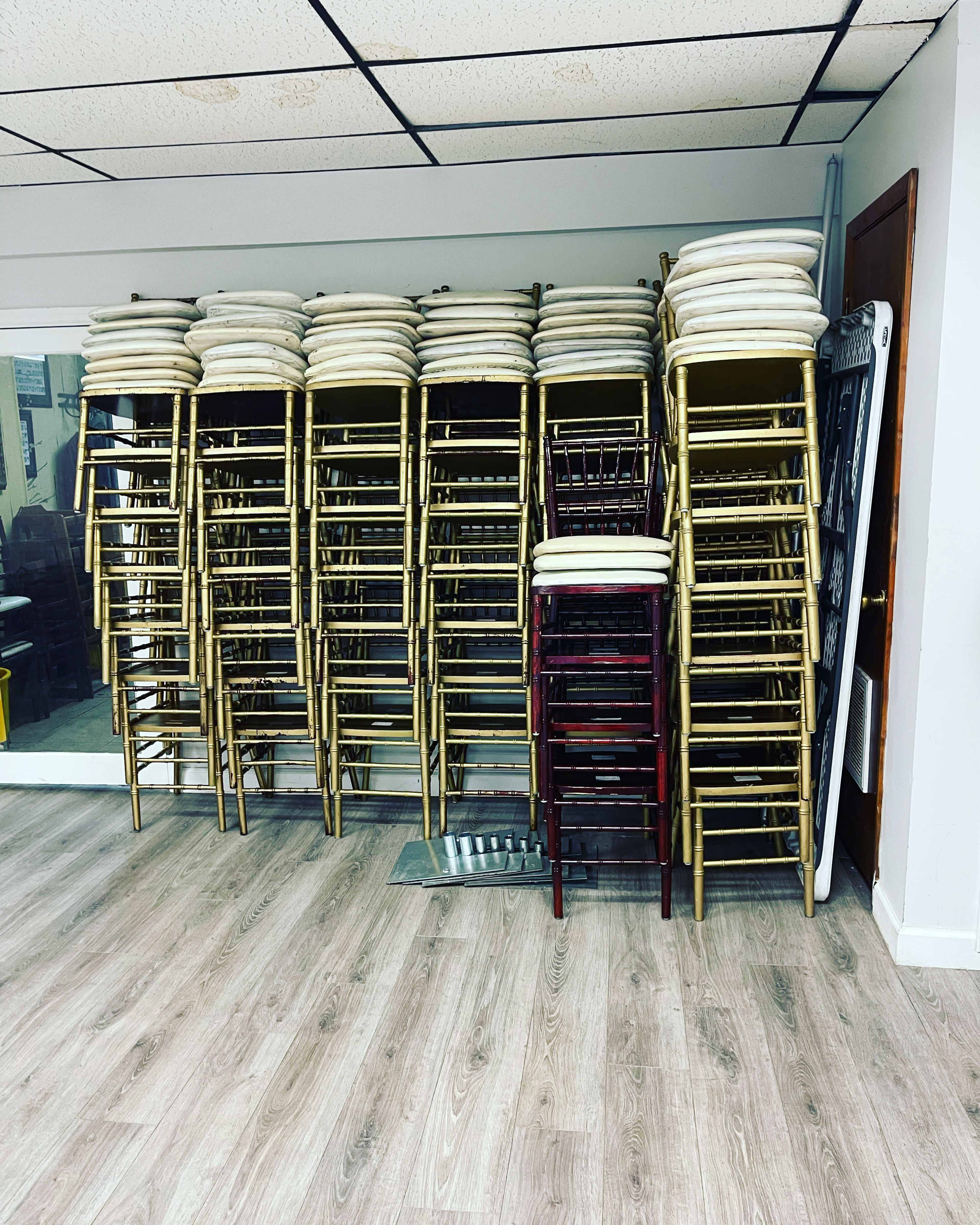 A stack of chairs with gold frames and white cushions is arranged against a wall in a room with wooden flooring.