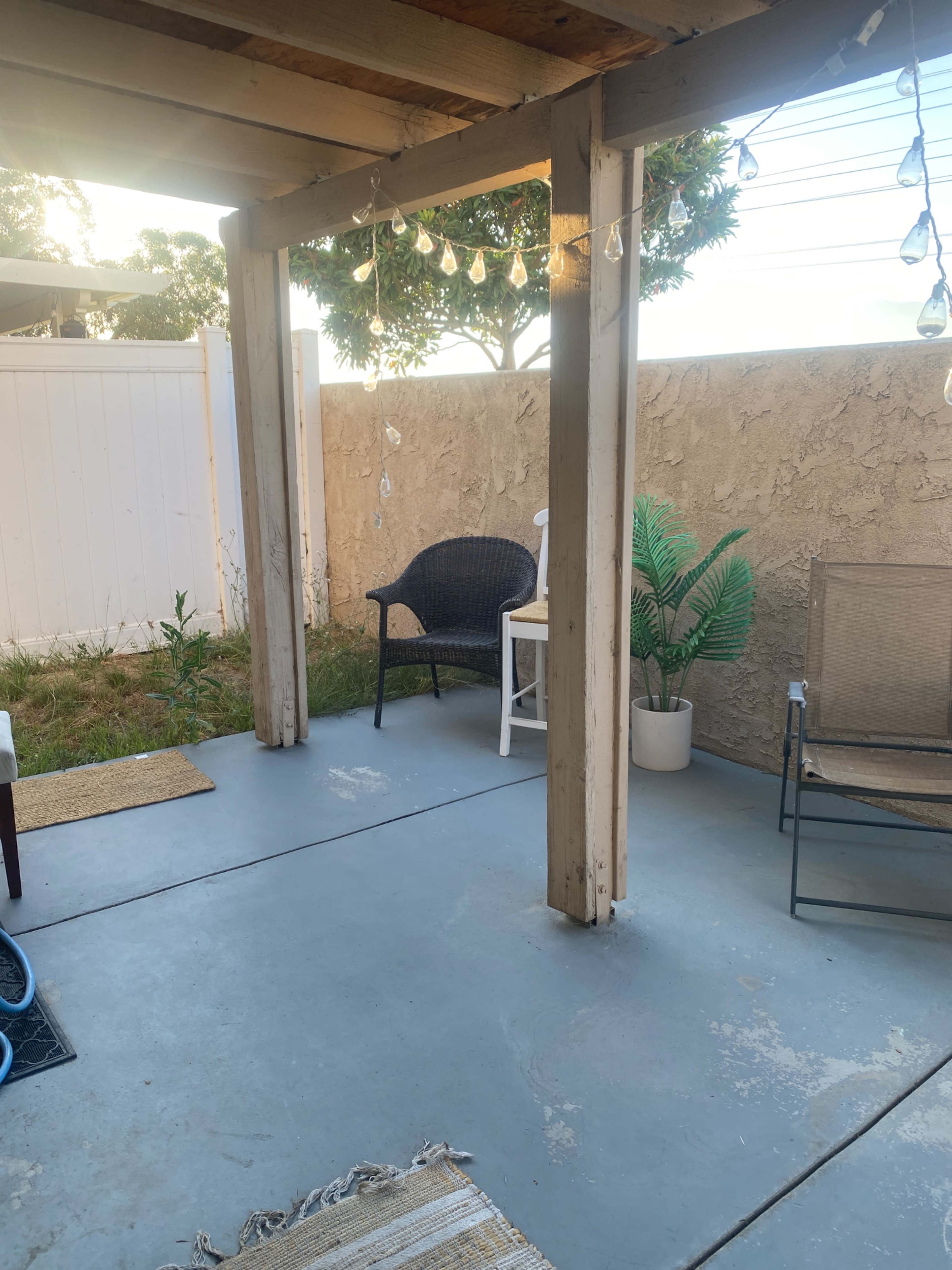 The image shows a small outdoor patio area with two chairs, a potted plant, and decorative string lights hanging from the overhead structure.