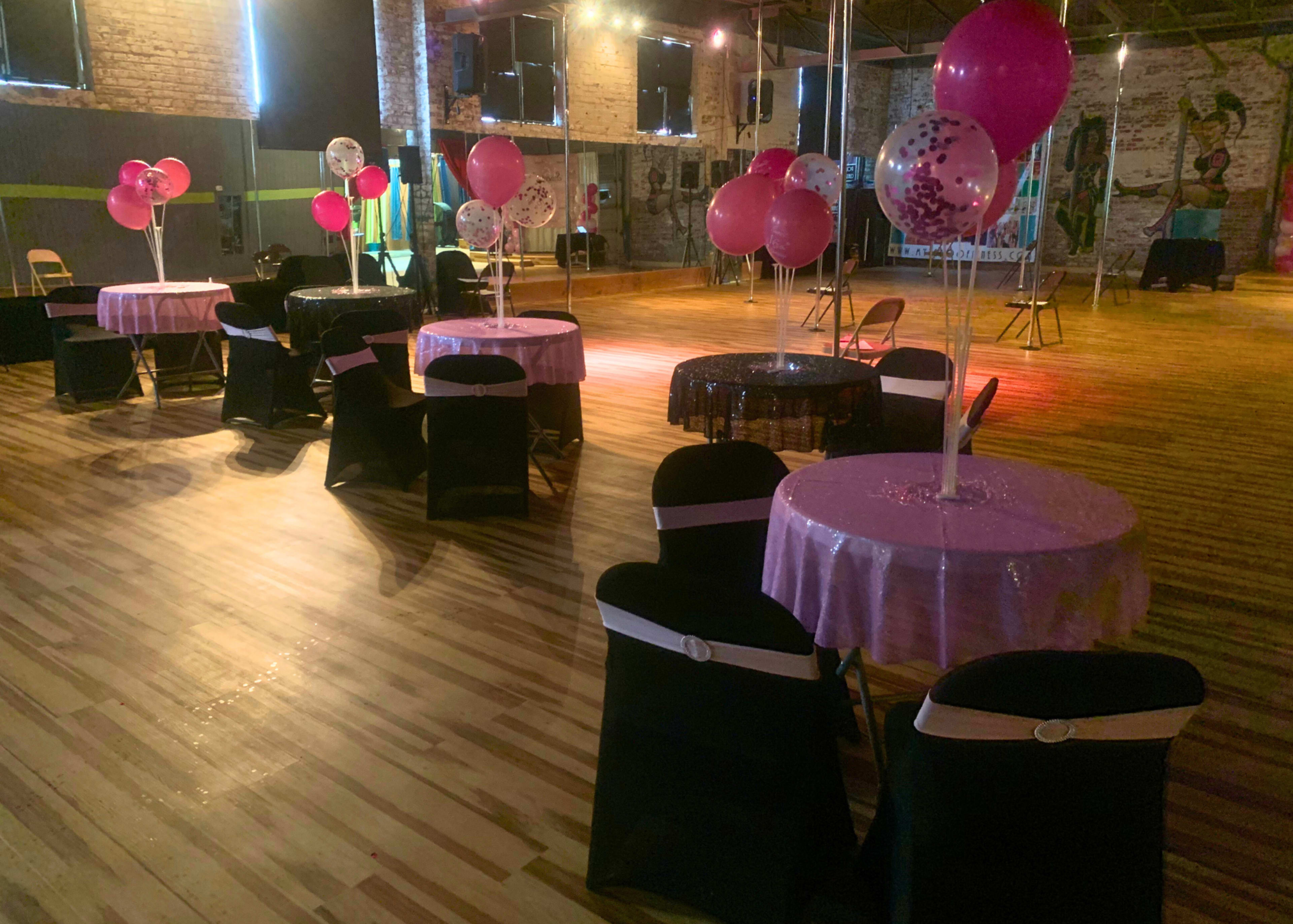 A spacious event venue with several tables covered in pink tablecloths, black chairs, and decorated with pink and clear balloons.