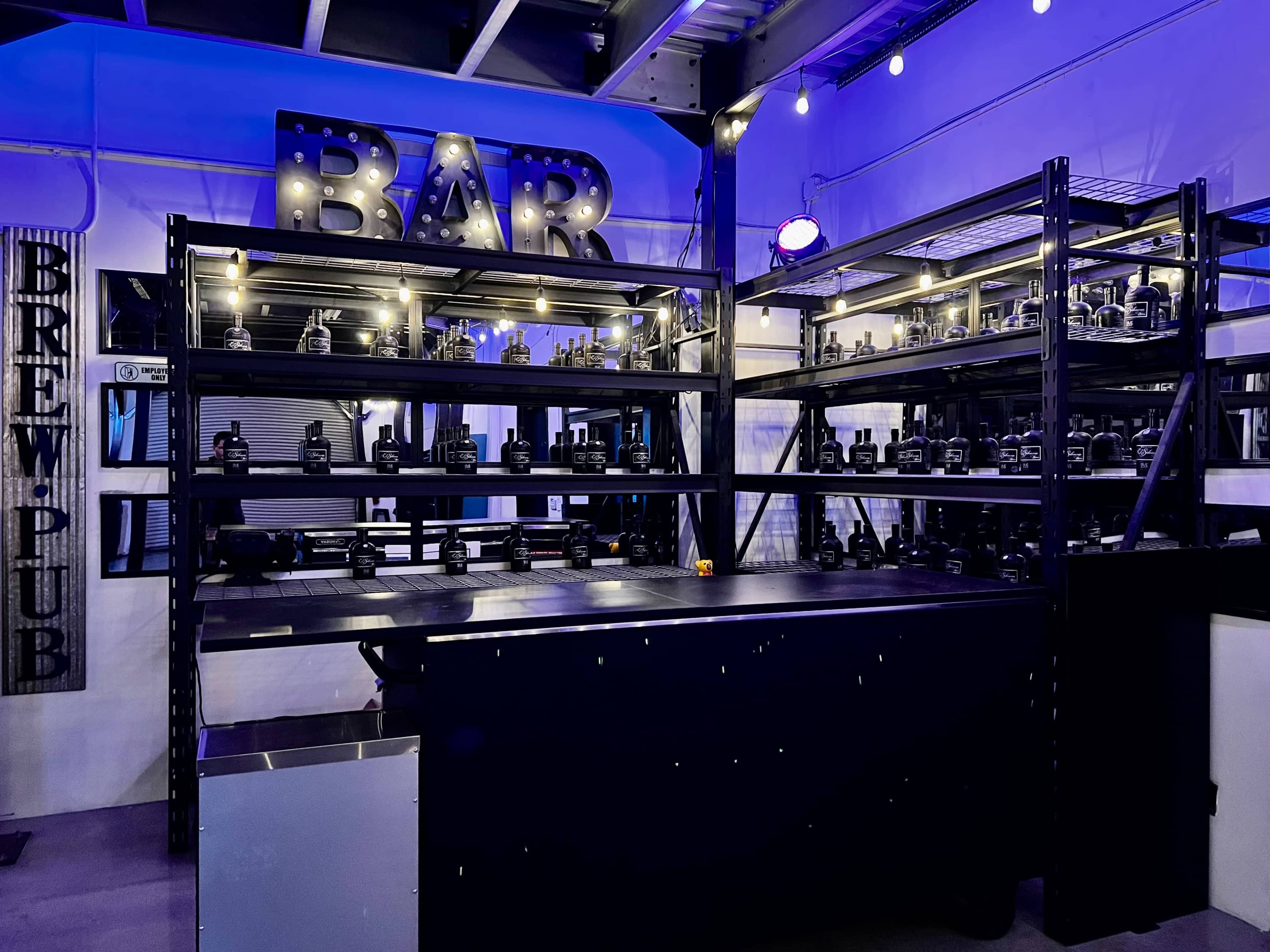 A bar area with shelves displaying various drinks, illuminated by purple lighting in a modern setting.