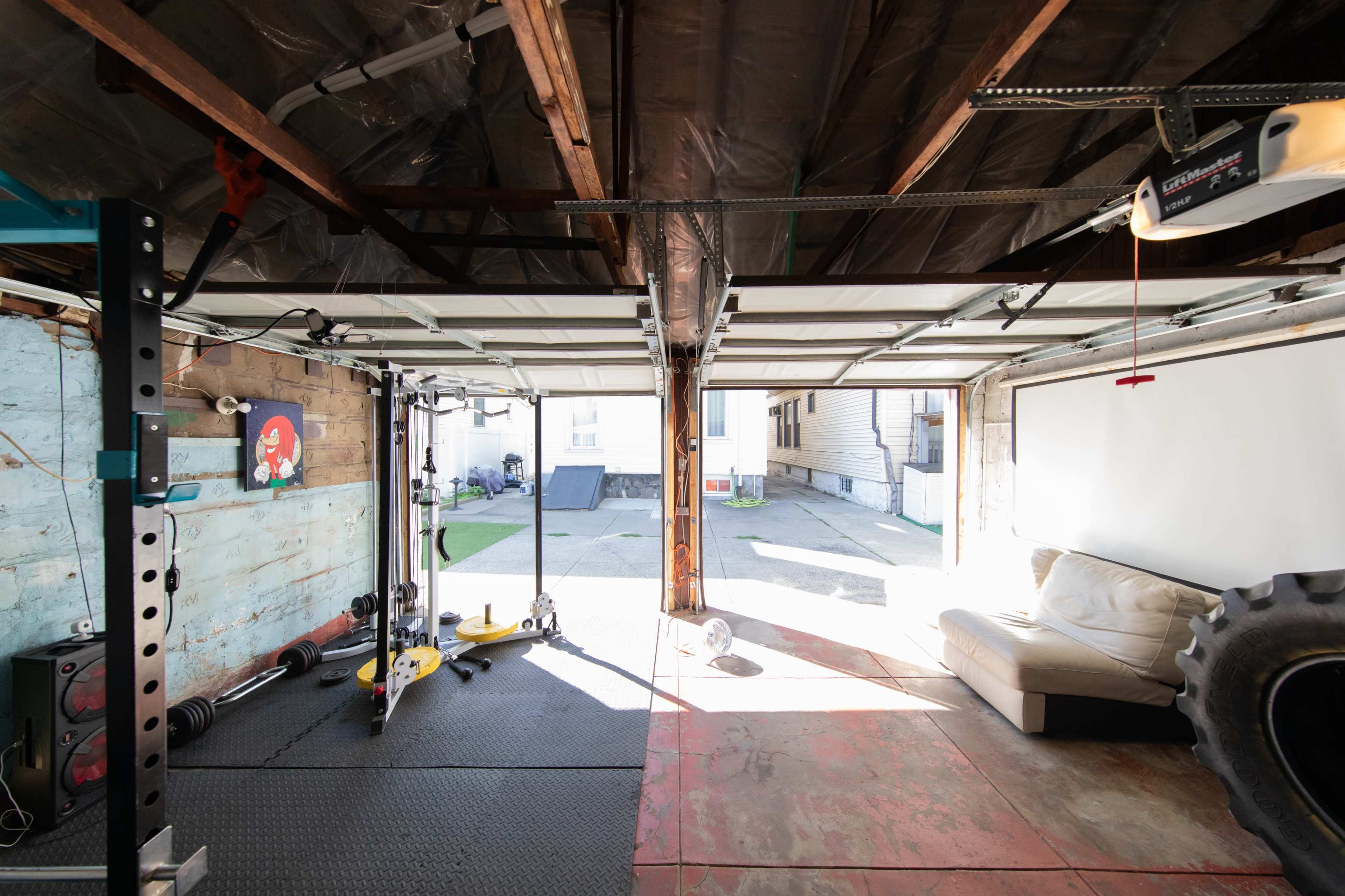 The image shows a spacious garage with an open door, revealing a workout area equipped with exercise equipment, a couch, and a tire in the corner.