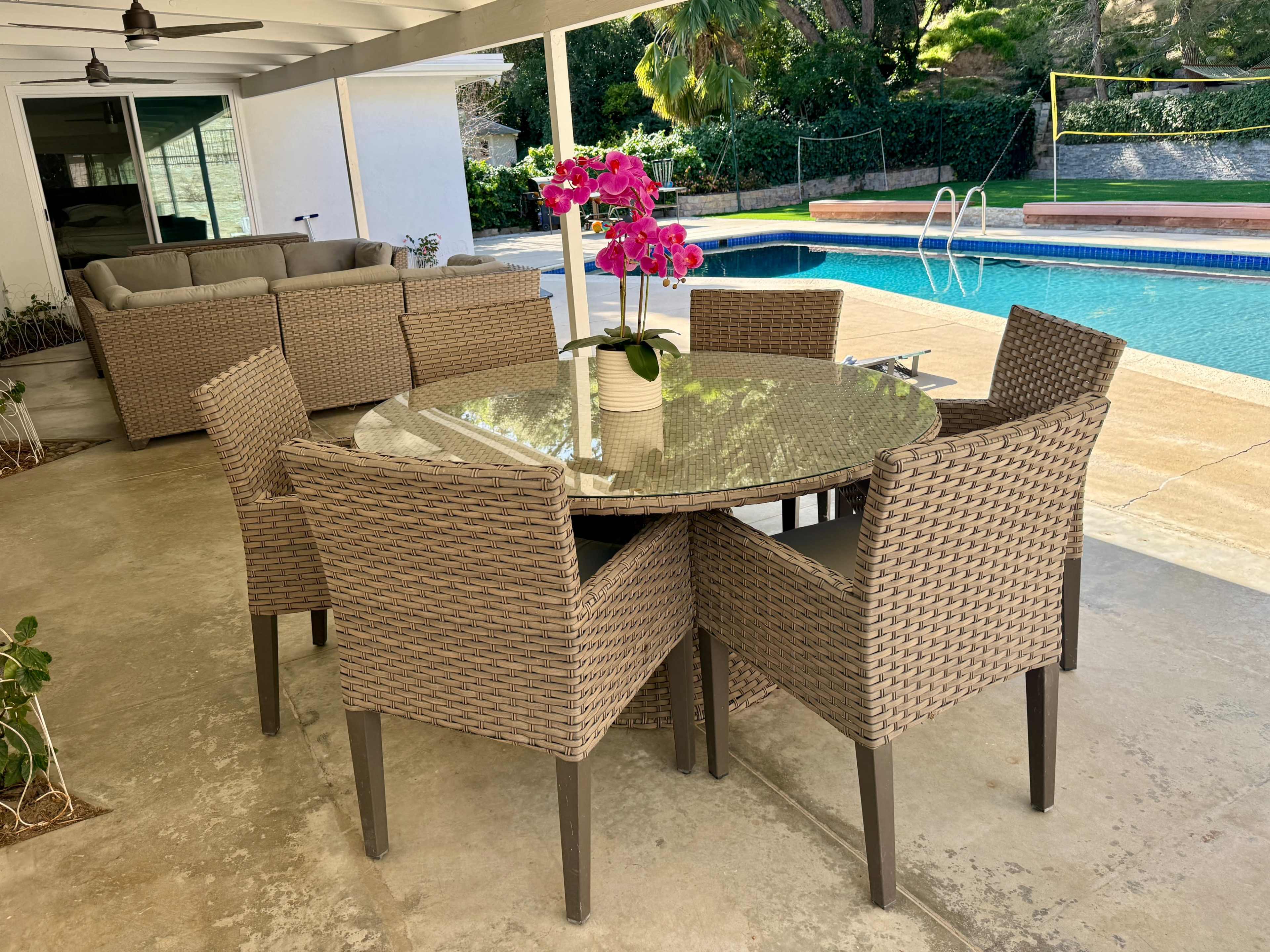 A round glass table with six wicker chairs is set under a covered patio next to a swimming pool surrounded by greenery.